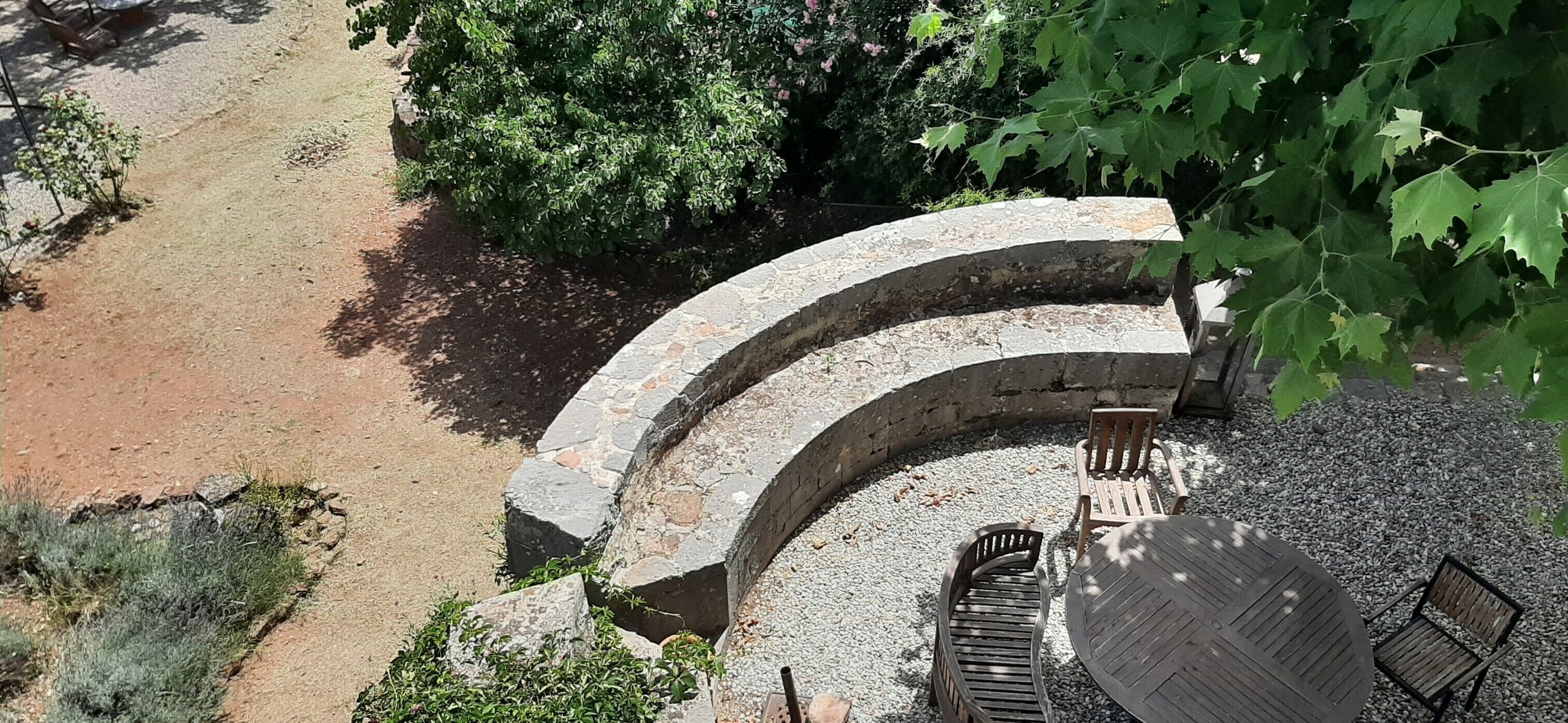 A stone bench and table set in a circular stone structure surrounded by gravel, with a dirt path and greenery in the background.