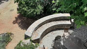 A stone bench and table set in a circular stone structure surrounded by gravel, with a dirt path and greenery in the background.