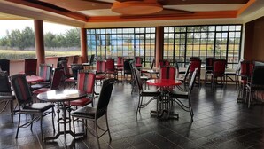 Restaurante com várias mesas e cadeiras, janelas amplas que oferecem vista para o exterior e piso ladrilhado.