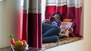 Uma pessoa lendo um livro chamado 'Harvard Business Review' em um quarto com janela, cortinas e uma tigela de frutas sobre uma mesa.