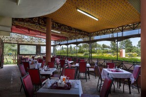 Un restaurante al aire libre con mesas y sillas, un área techada con techo estampado y vistas a un jardín.