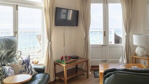 A living room with a view of the sea, a television, a table with a board game, and a small sailboat model. - Garden Apartment On The Beach (Totland Bay)