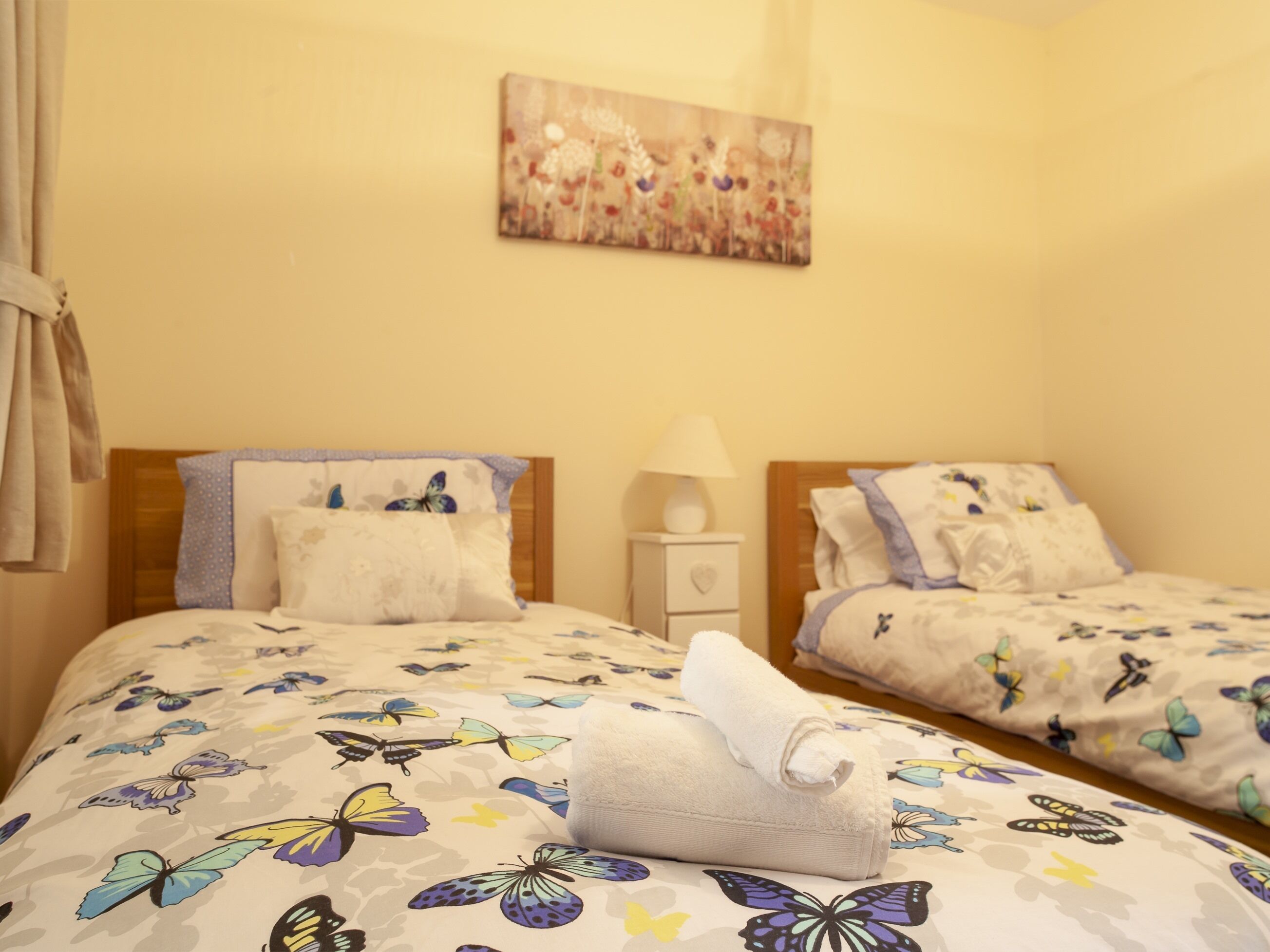 A hotel room with two beds, each with a floral and butterfly-themed bedspread. There is a painting on the wall above the beds and a bedside table with a lamp.