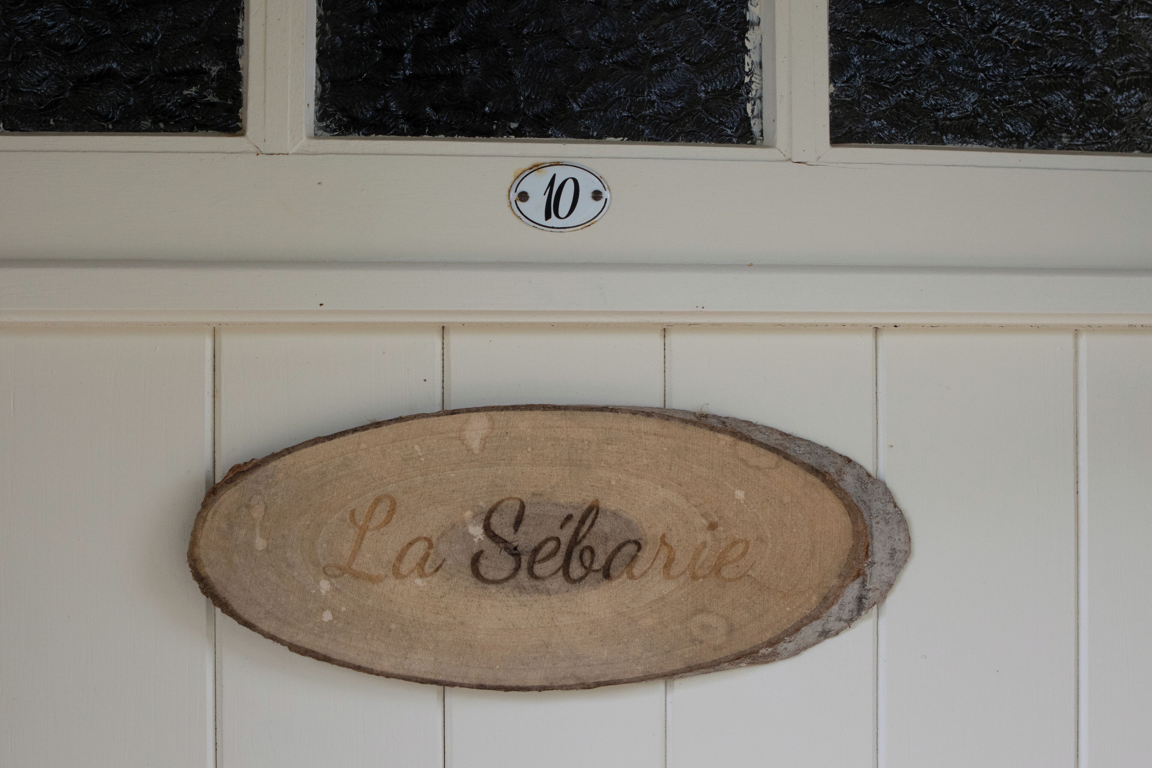 Un letrero de madera con el nombre "La Sébarie" grabado, montado en una pared blanca sobre un ventanal con un marco negro con textura.