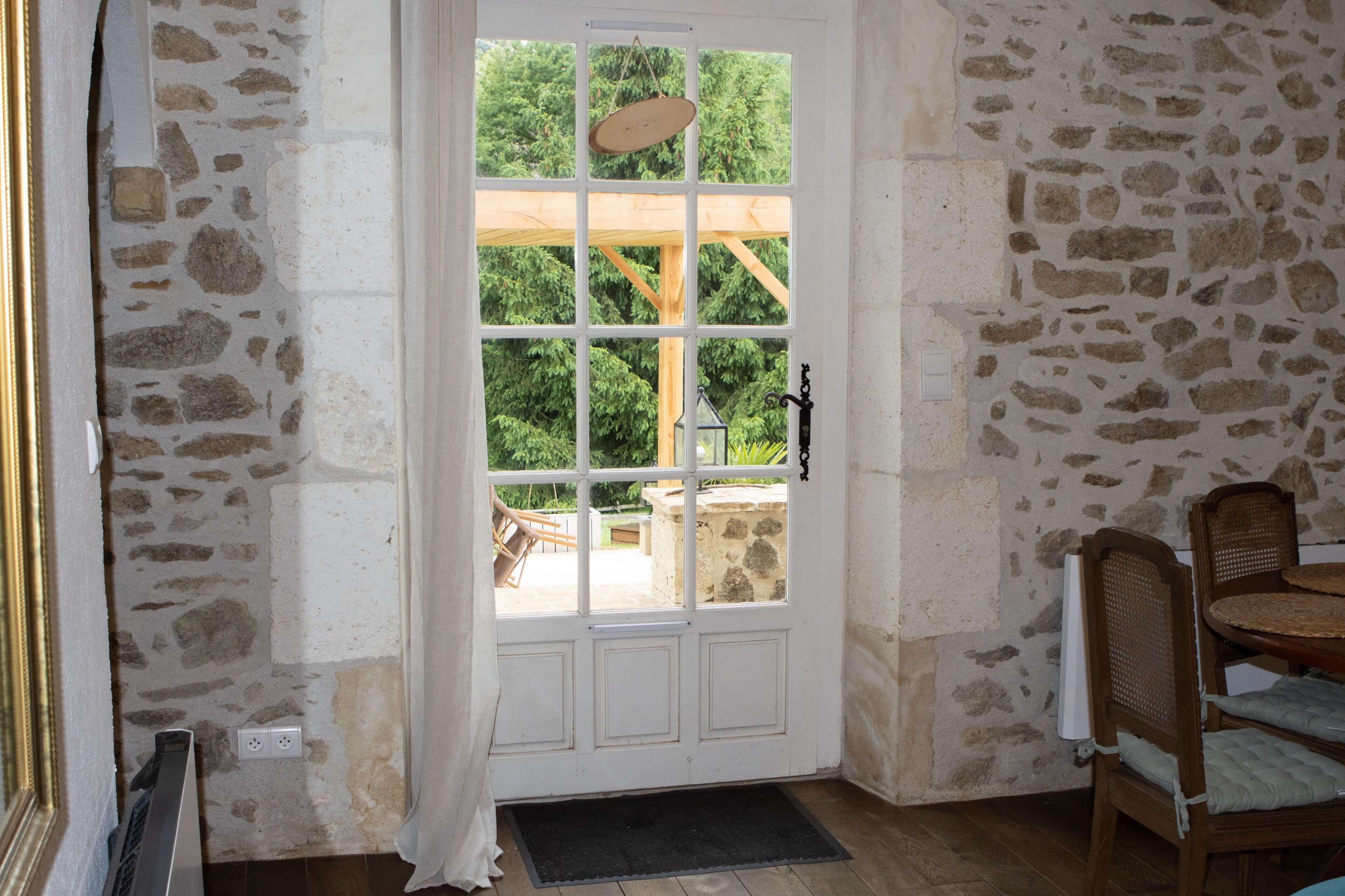 Une pièce aux murs de pierre, avec une porte blanche, une petite table entourée de chaises et une fenêtre donnant sur un paysage verdoyant.