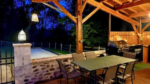 Un patio al aire libre con un muro de piedra, una pérgola de madera, una parrilla y una mesa con sillas.