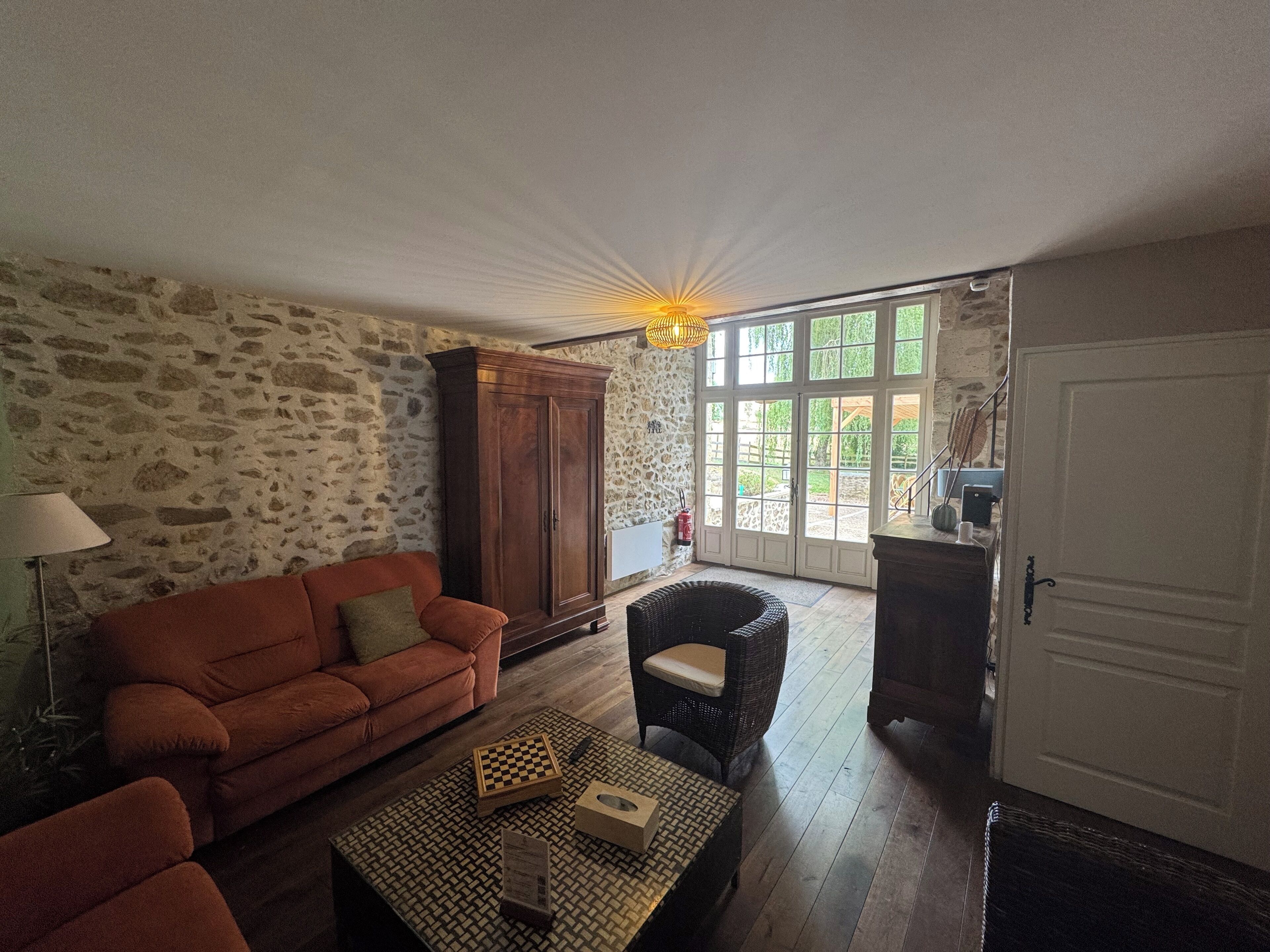 A cozy living room with a stone wall, a wooden cabinet, a brown sofa, a wicker chair, and a coffee table with a chessboard.