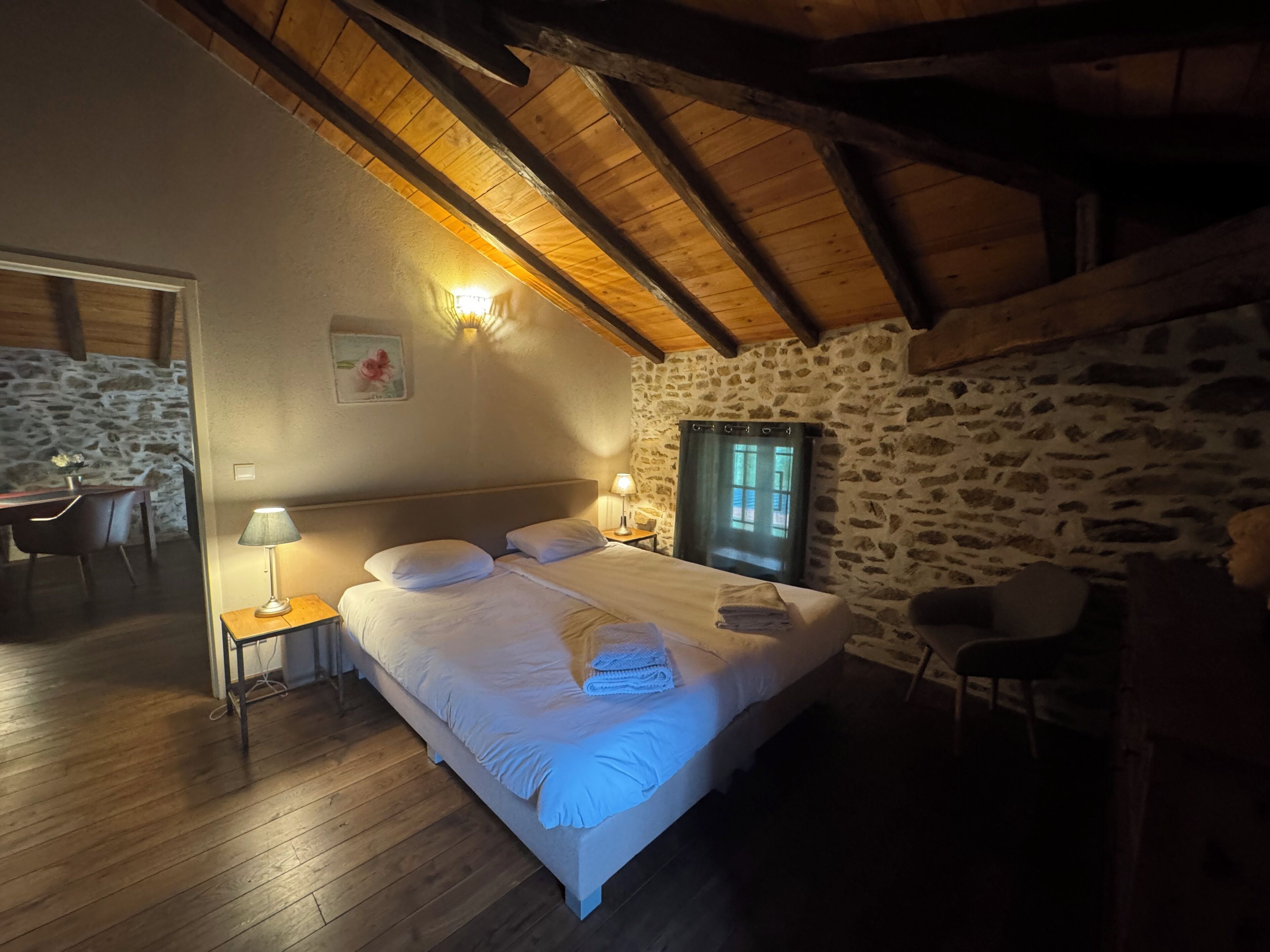 Un dormitorio acogedor con pared de piedra, vigas de madera en el techo, cama con ropa blanca y mesita de noche con lámpara.