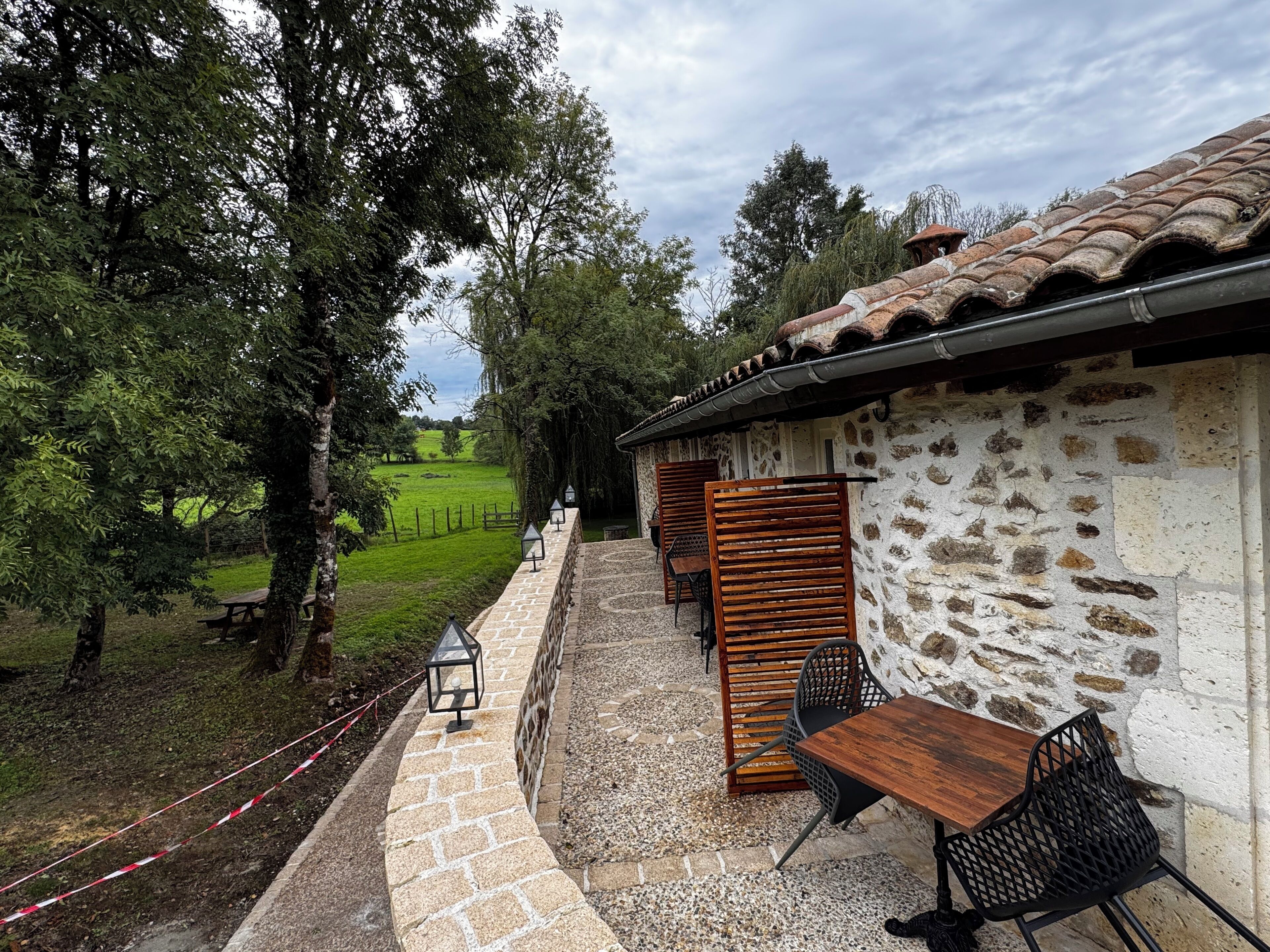 A stone building with a tiled roof, outdoor seating with a table and chairs, and a view of a green field and trees.