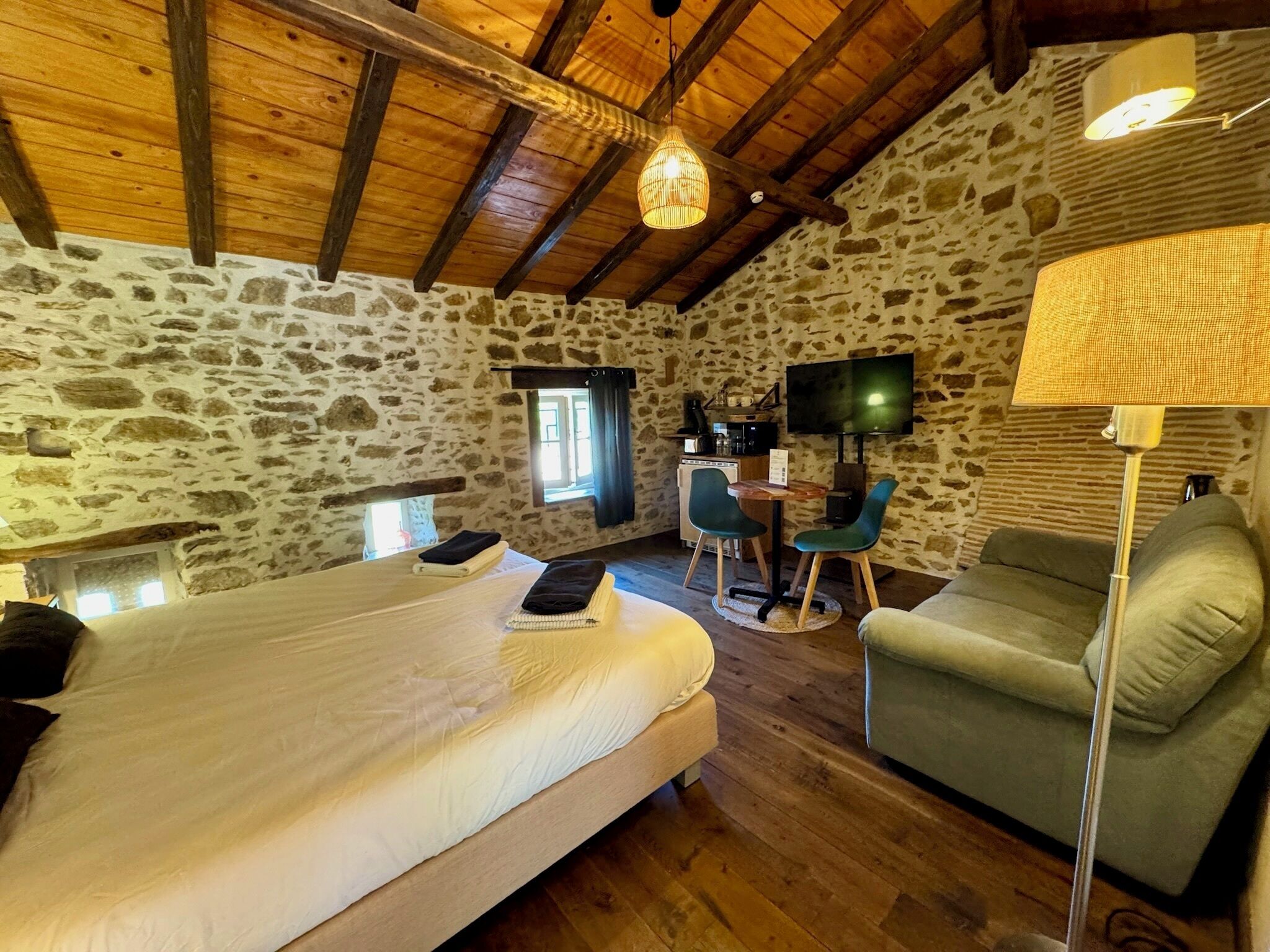 Une pièce confortable avec un lit, un fauteuil vert, un bureau avec une chaise, une télévision et un mur en pierre.