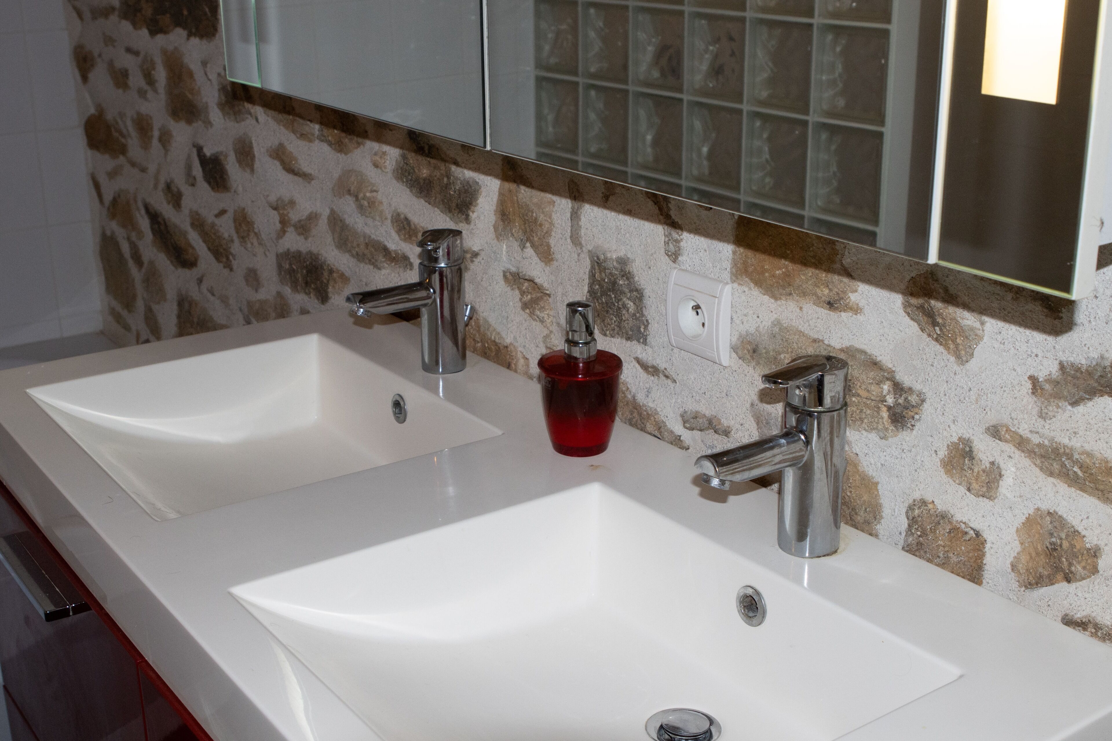 Une salle de bain avec deux lavabos blancs, un distributeur de savon rouge et un miroir au-dessus.