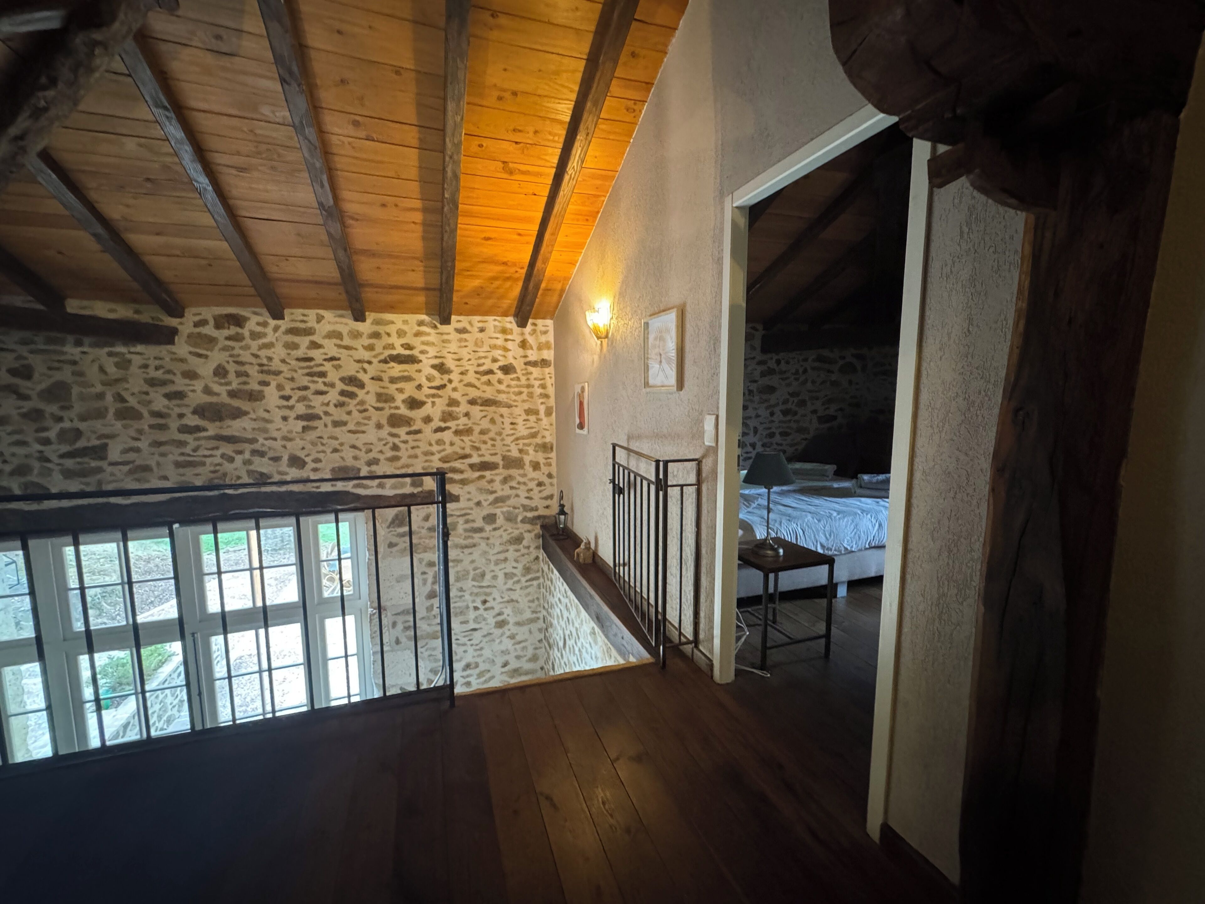 A rustic room with stone walls, wooden beams, a bed, and a staircase leading to an upper level.