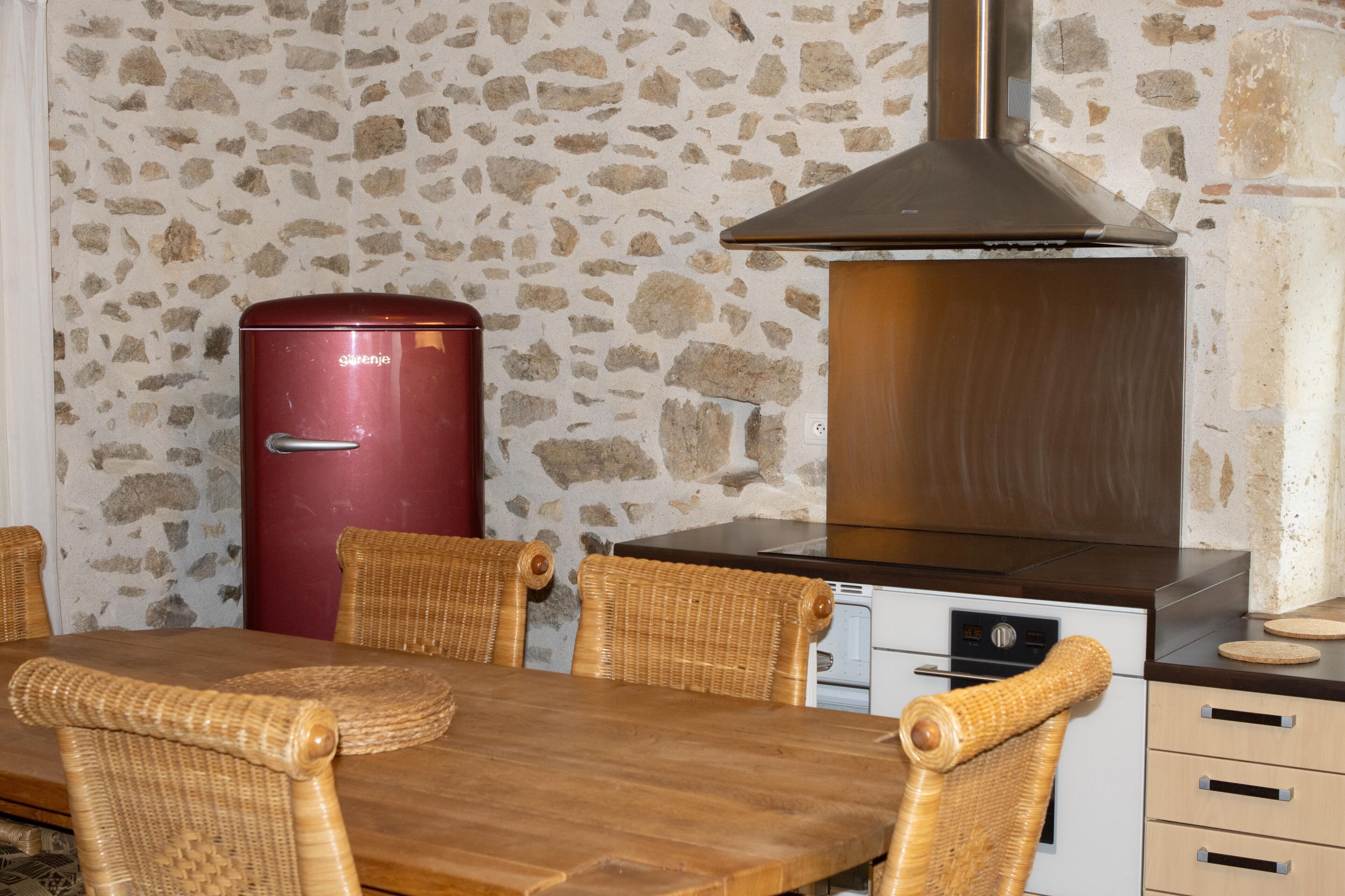 A dining area with a stone wall, a red appliance, a stainless steel range hood, wicker chairs, and a wooden table.