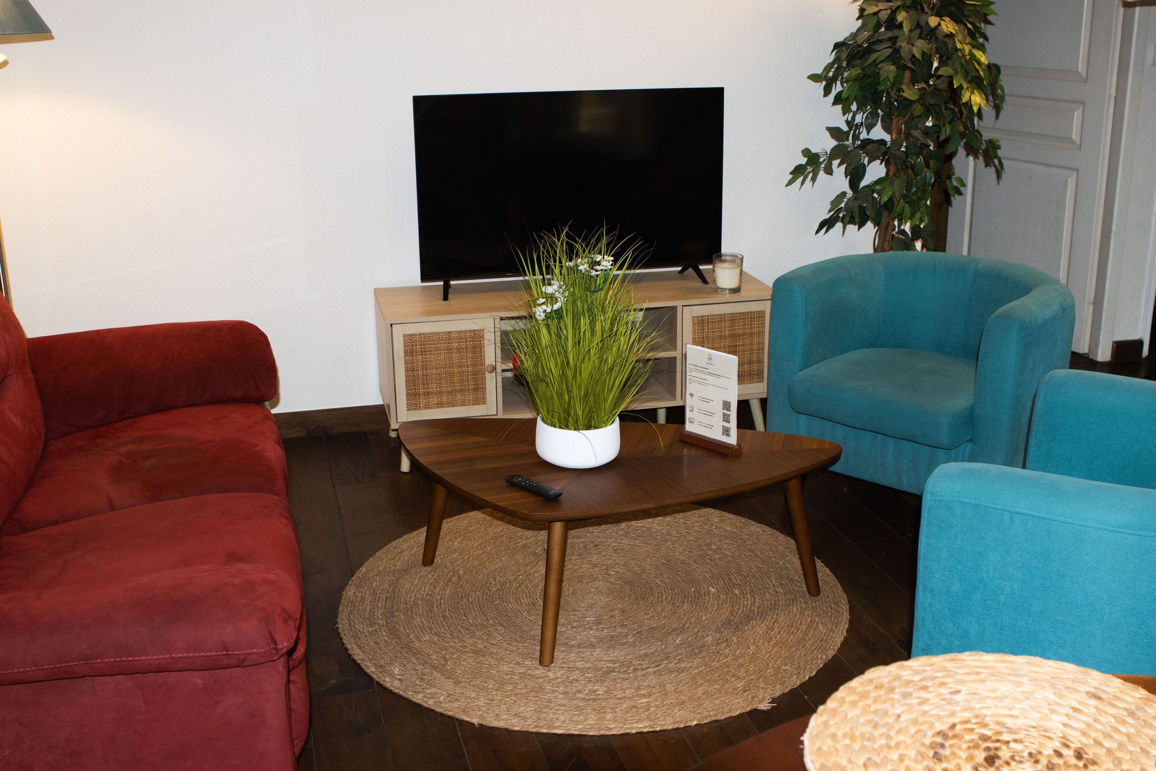 A living room with a flat-screen TV, a red sofa, a blue armchair, and a small potted plant on a wooden coffee table.