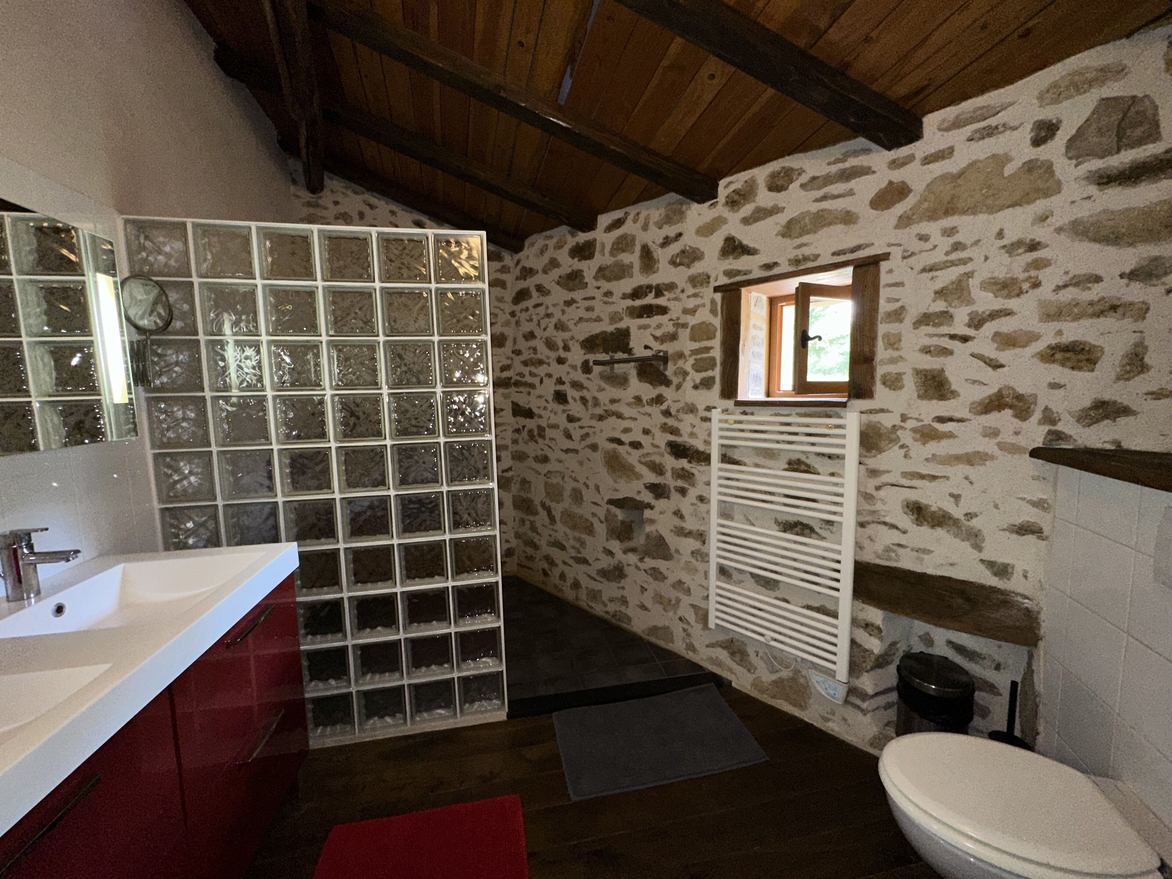 A bathroom with a stone wall, a wooden ceiling, a glass shower enclosure, a white sink, and a toilet.