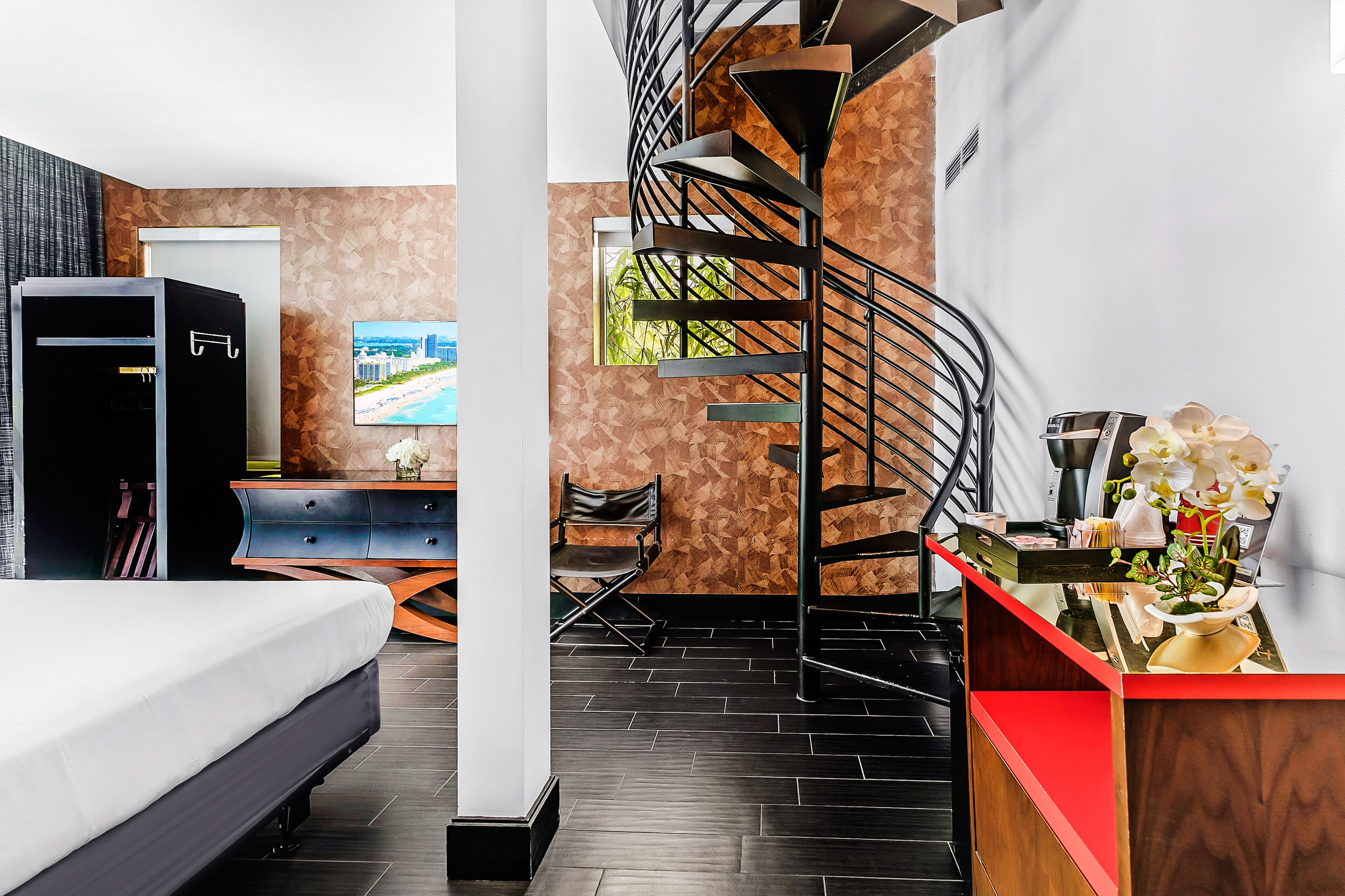 A modern hotel room with a bed, a desk with a computer, a TV, a spiral staircase, and a view of the outdoors.