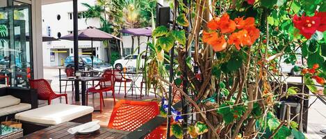 An outdoor cafe with tables and chairs, surrounded by lush greenery and flowering plants.