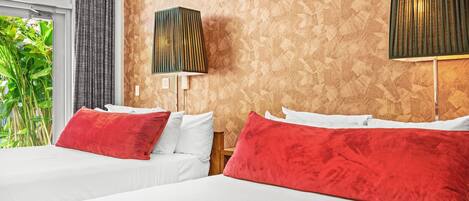 A hotel room with two beds, red and white bedding, a wooden textured wall, and a window with a view of greenery.