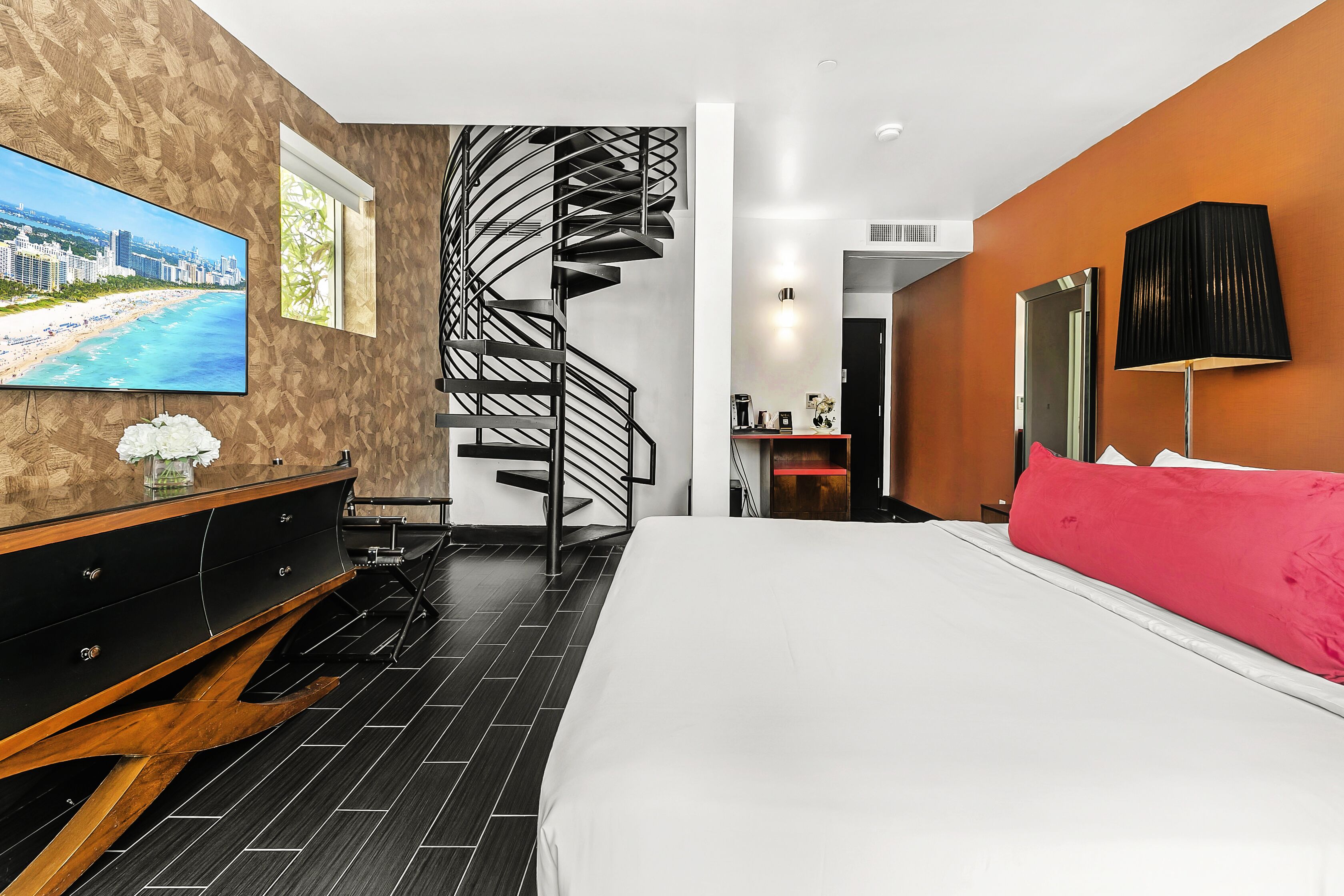A modern hotel room with a large bed, a TV on the wall, a wooden dresser, and a spiral staircase.