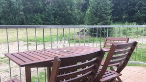 Ein Holztisch mit Stühlen auf einem Balkon, der von einem Metallgeländer gesäumt wird und auf eine begrünte Fläche mit Bäumen blickt.