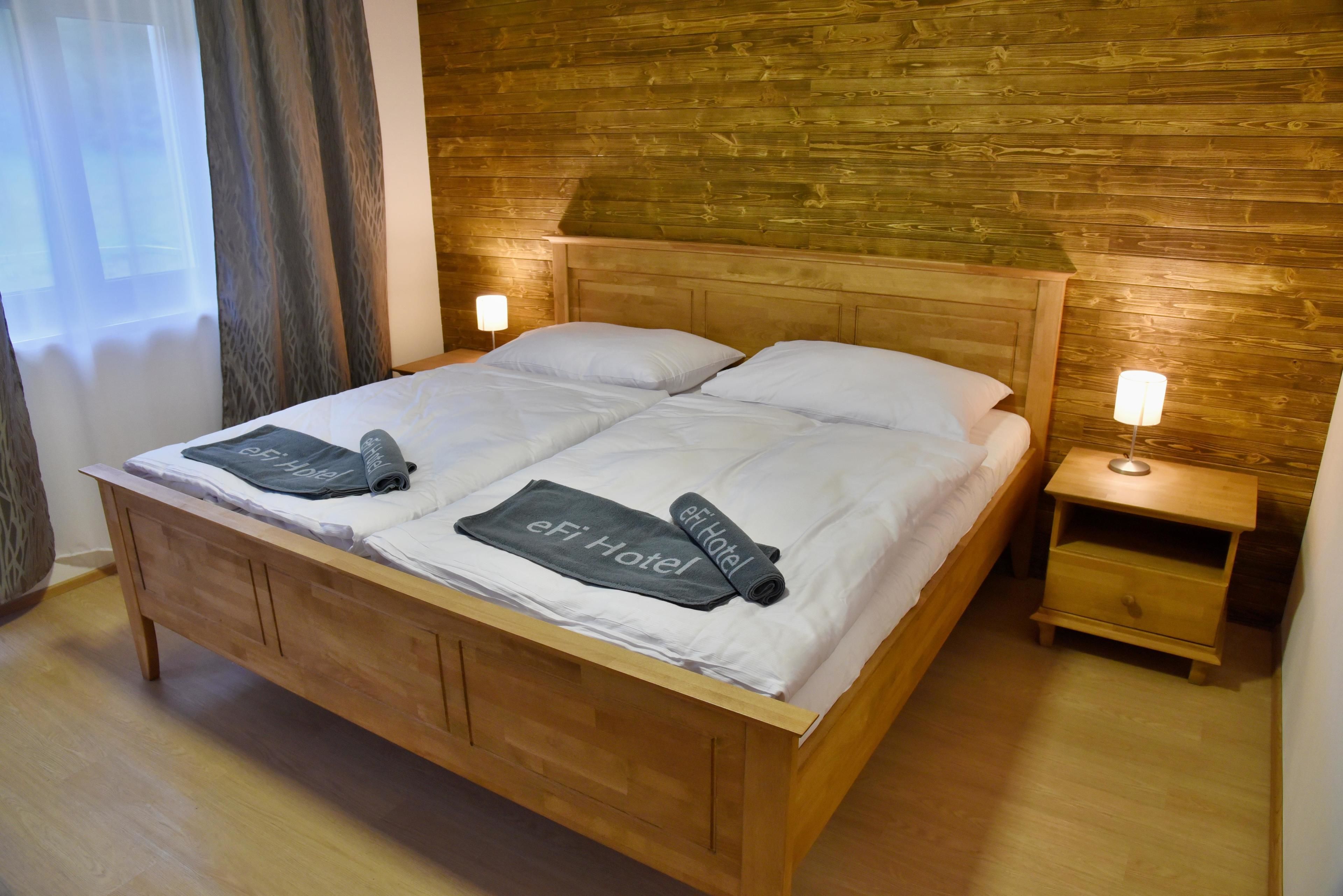 Una cama de madera con dos almohadas y una colcha, una mesita de noche con una lámpara, y un cabecero de madera.
