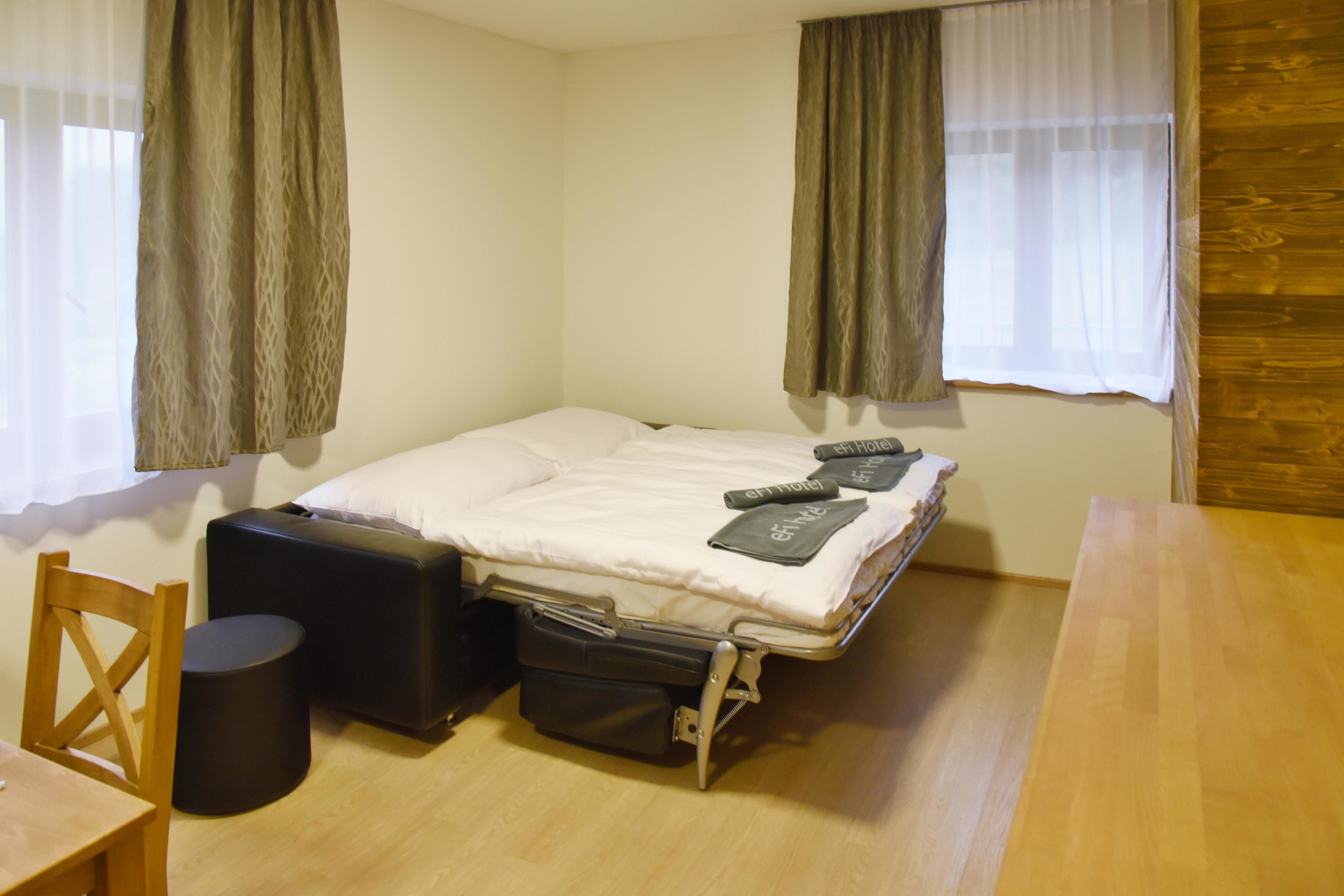 A small, compact room with a single bed, a wooden table, a chair, and a suitcase.