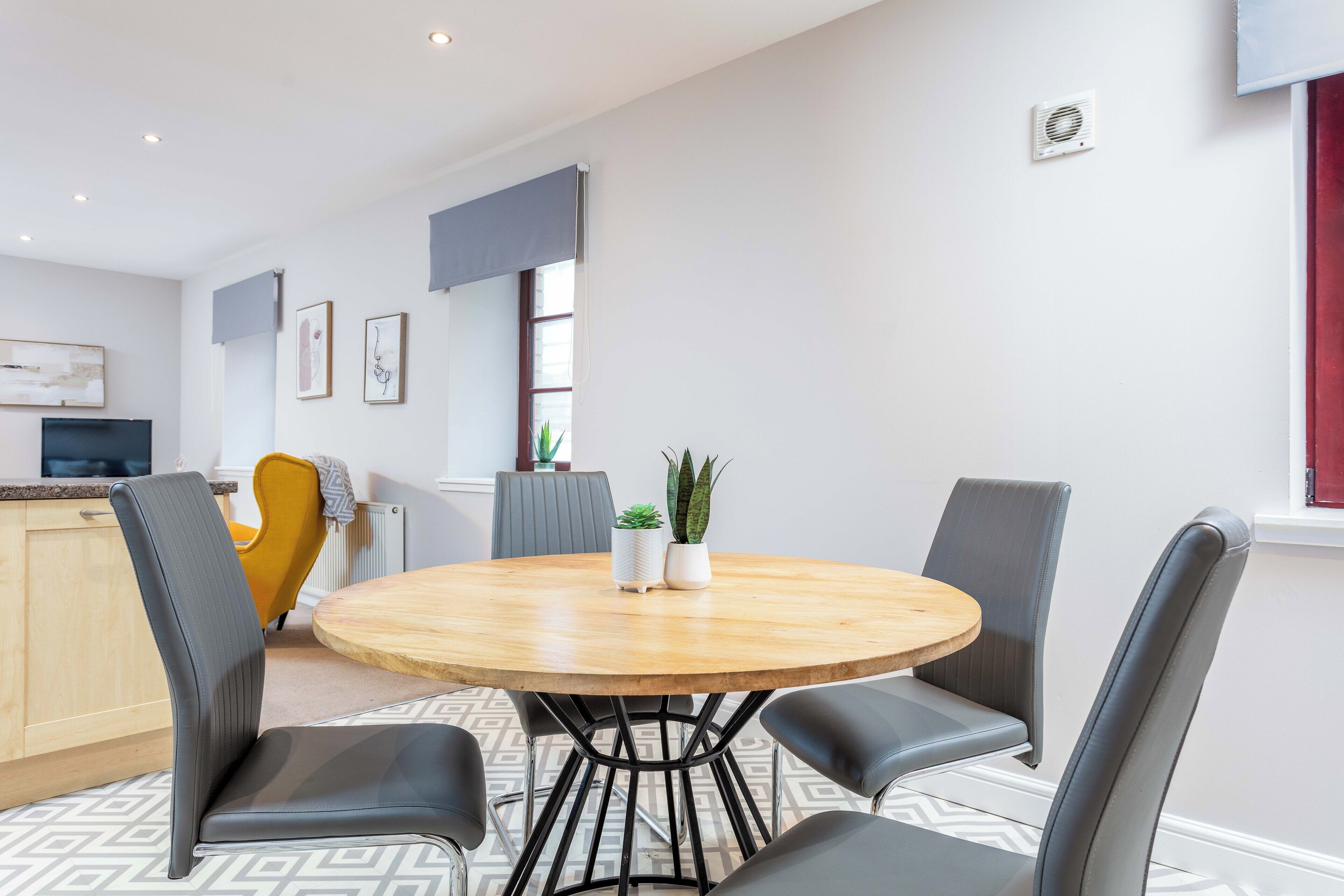 A modern kitchen with a round wooden table, grey upholstered chairs, and a yellow chair. There are potted plants on the table and a radiator in the background.