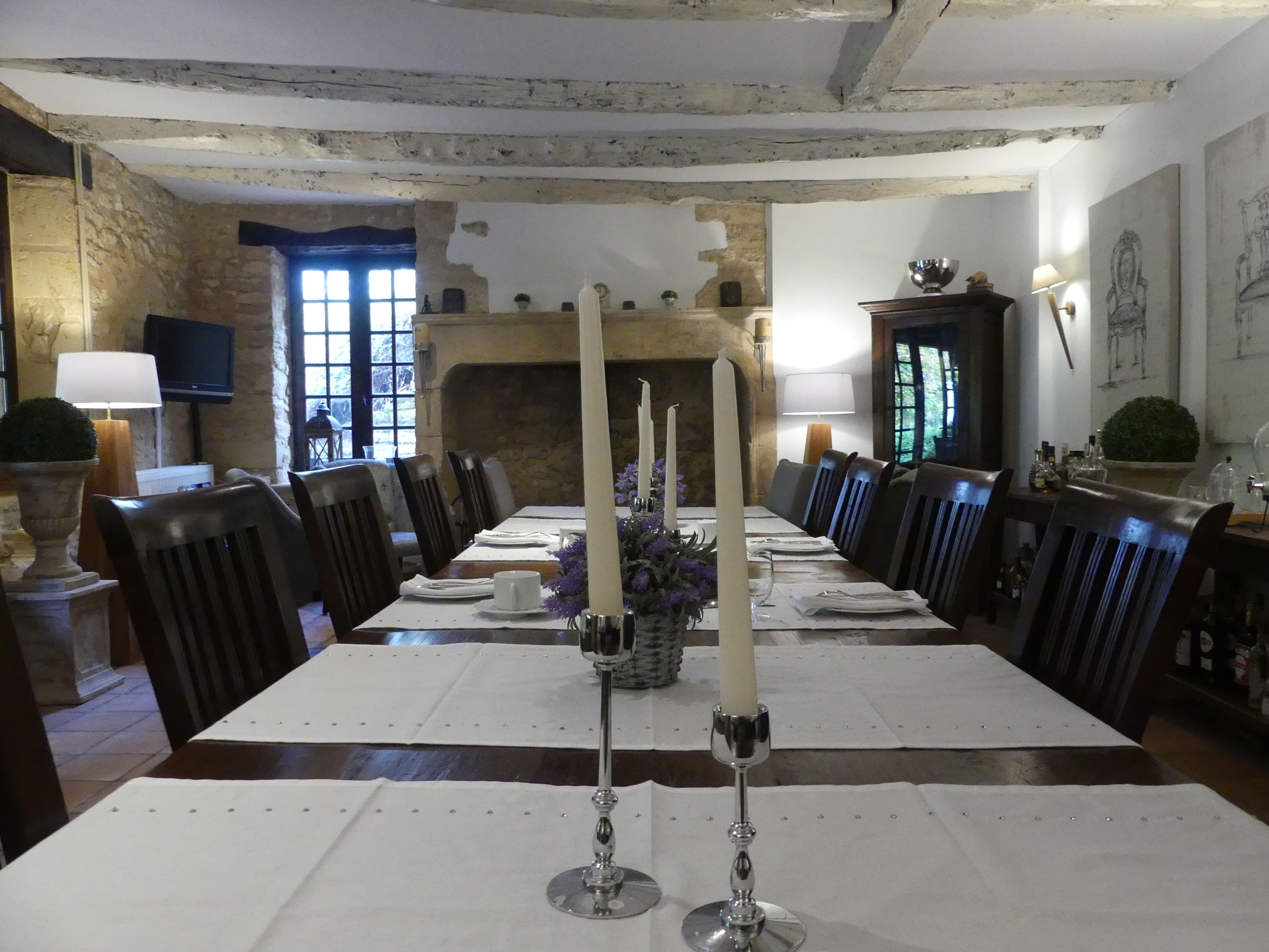 A dining room with a long table set for a meal, featuring candles, a vase with flowers, and a fireplace in the background.