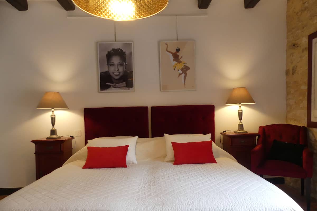 A bed with a red headboard, white bedding, and two red pillows. There are two bedside lamps, a red chair, and two framed pictures on the wall.
