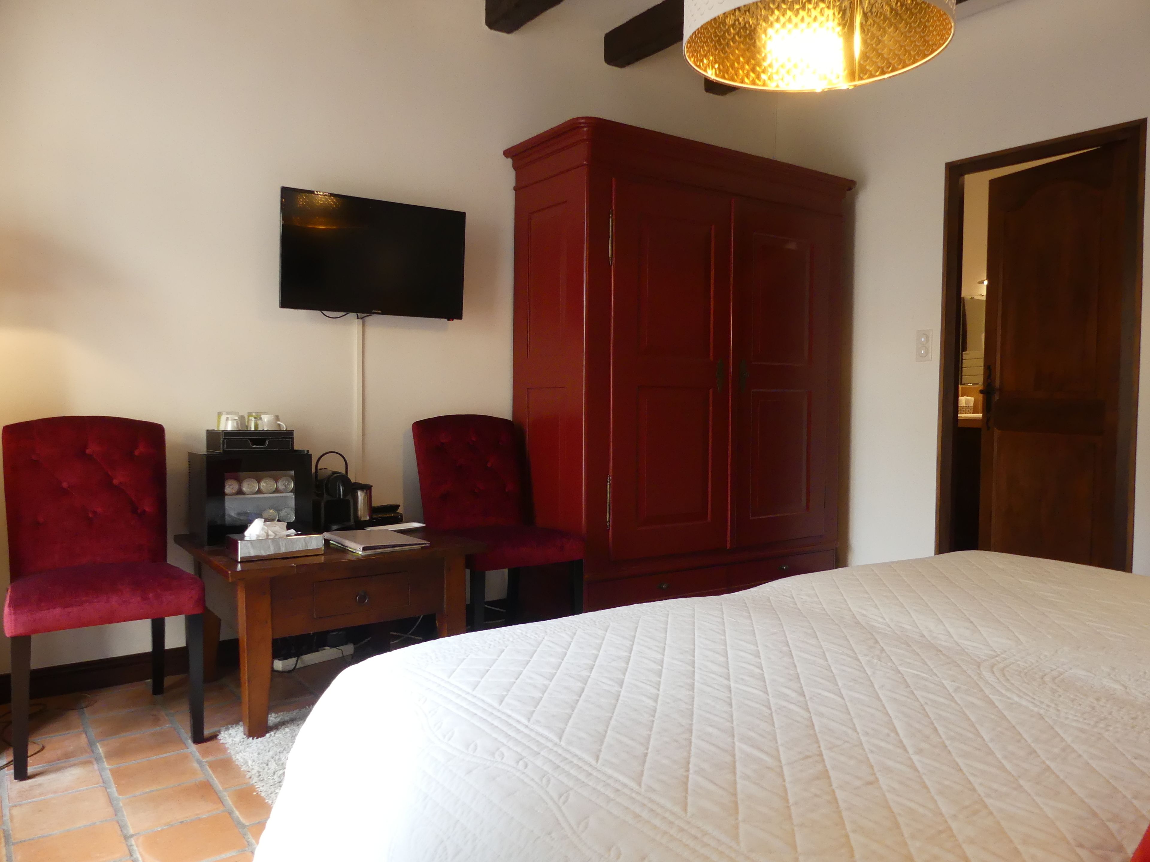 A bedroom with a large bed, two red upholstered chairs, a wooden nightstand, a television, and a red wardrobe.