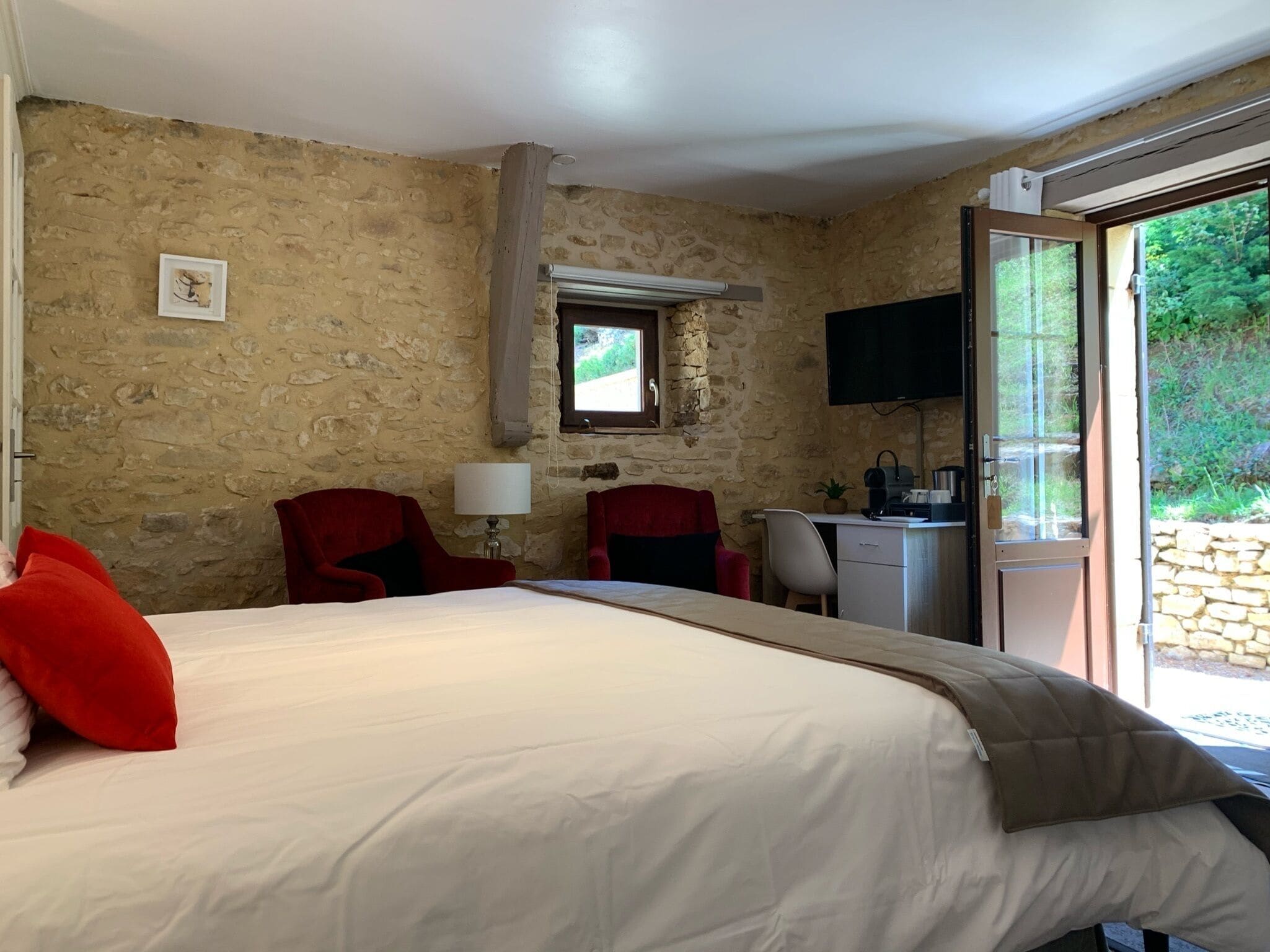 Un dormitorio con una cama grande, dos sillas rojas, un escritorio con una computadora, un televisor y una ventana con vistas a la vegetación.