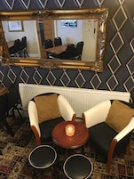 Een kamer met een spiegelwand, een radiator, twee fauteuils, een klein tafeltje met een kaars en een kamerbreed tapijt met een patroon.