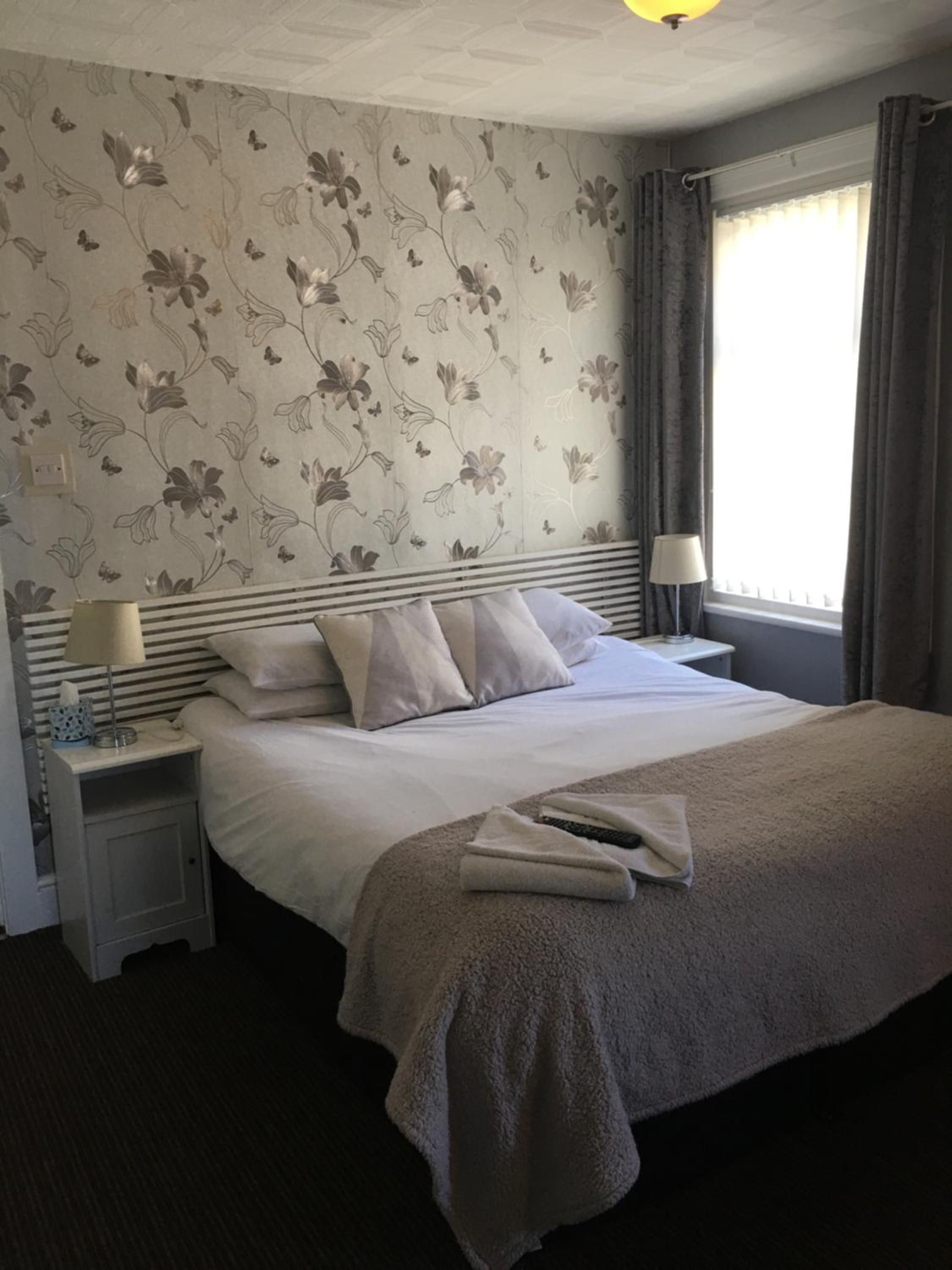 A hotel room with a bed, two bedside lamps, a window with curtains, and a floral wallpaper design.
