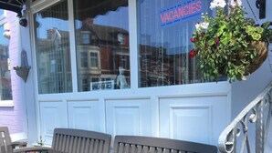 An outdoor seating area with a bench, a hanging flower pot, and a window with a 'VACANCIES' sign. - Glenheath Hotel (Blackpool)