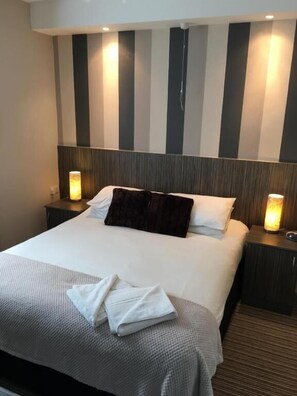 A neatly made bed with a dark throw pillow, two bedside lamps, and a striped wall behind. - Glenheath Hotel (Blackpool)