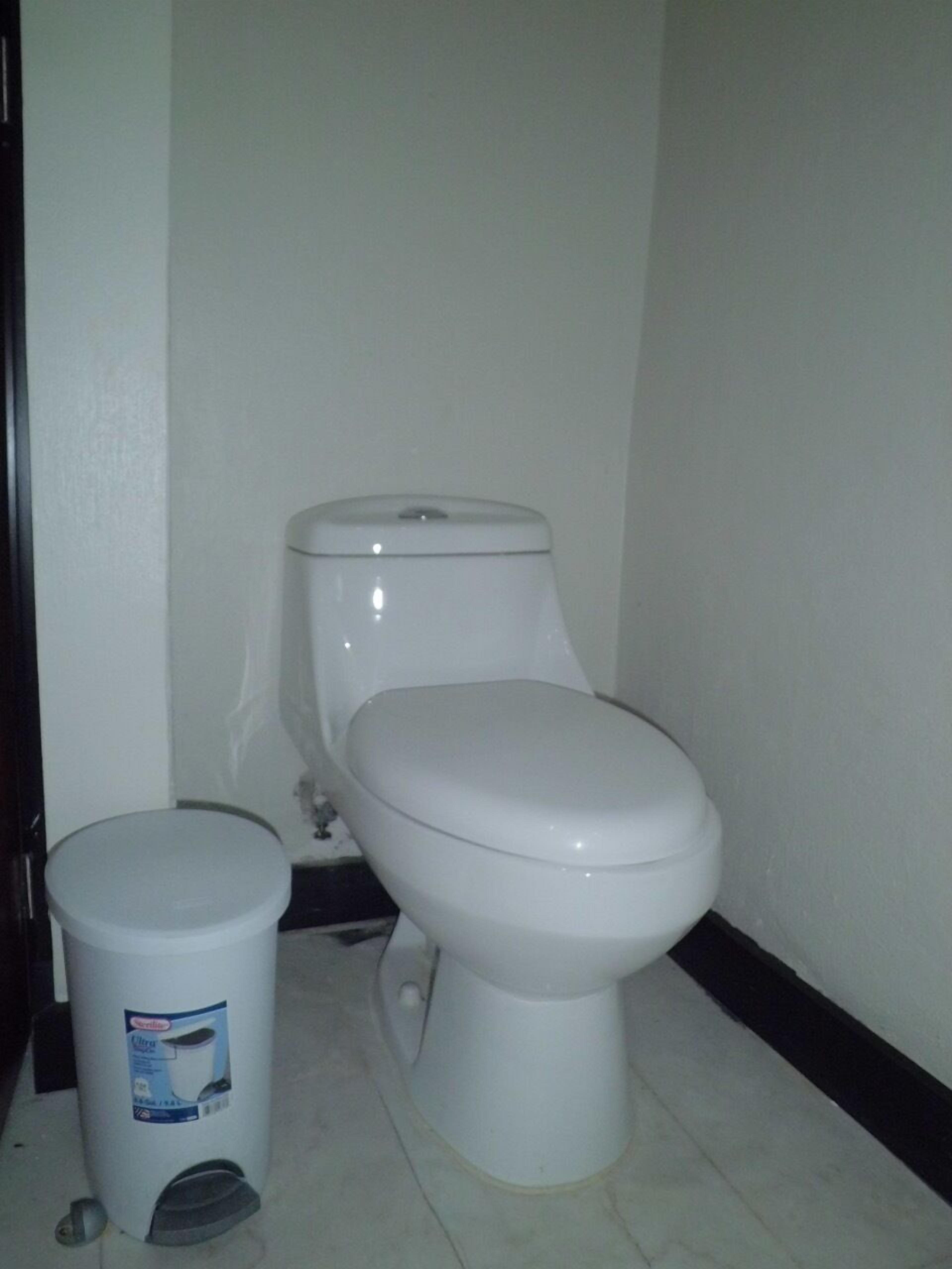 Dans une salle de bains aux murs blancs et au sol carrelé, on trouve des toilettes et une poubelle.