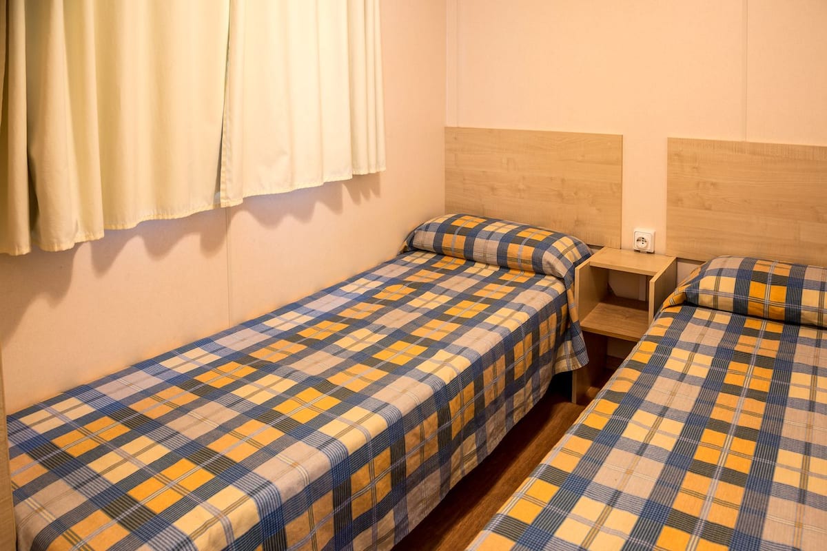 A small, simple room with two single beds, each with a plaid bedspread. There is a wooden headboard on one side and a white wall on the other. A small wooden nightstand is between the beds.
