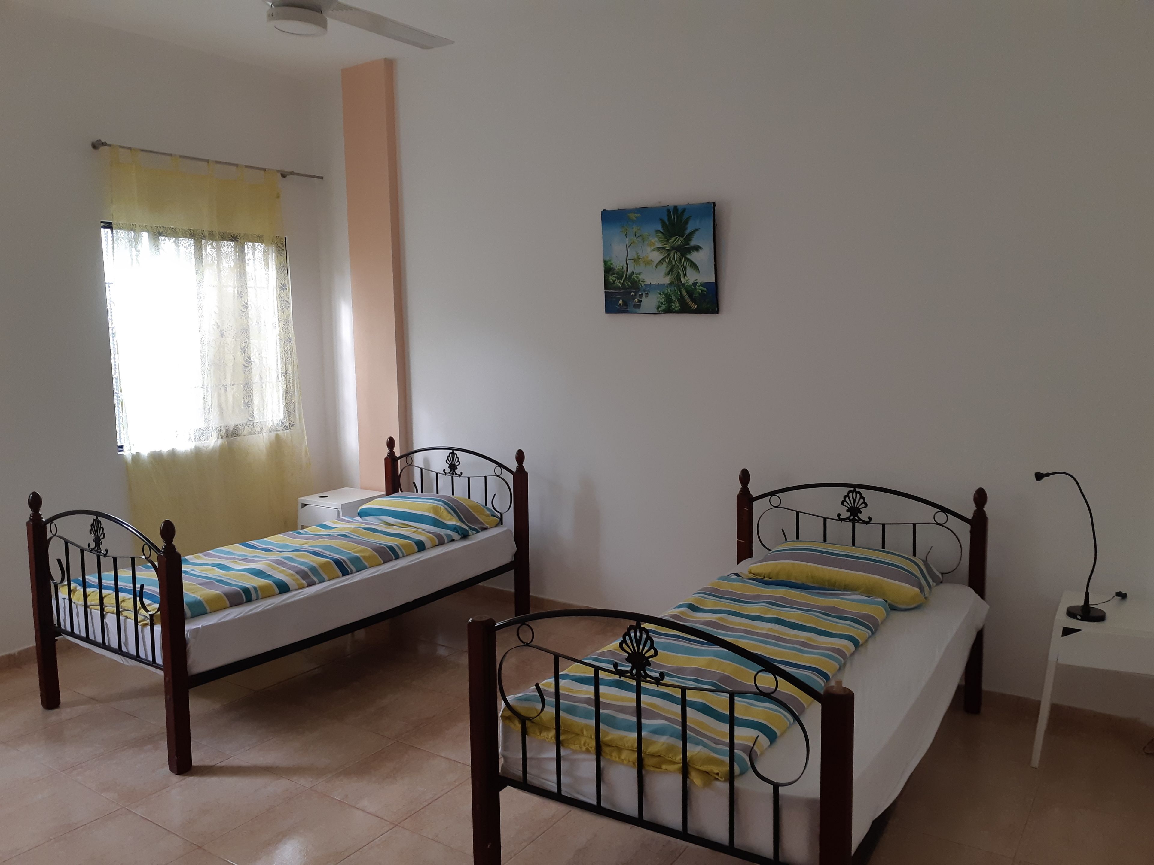 A room with two single beds, a window with curtains, a painting on the wall, and a nightstand with a lamp.