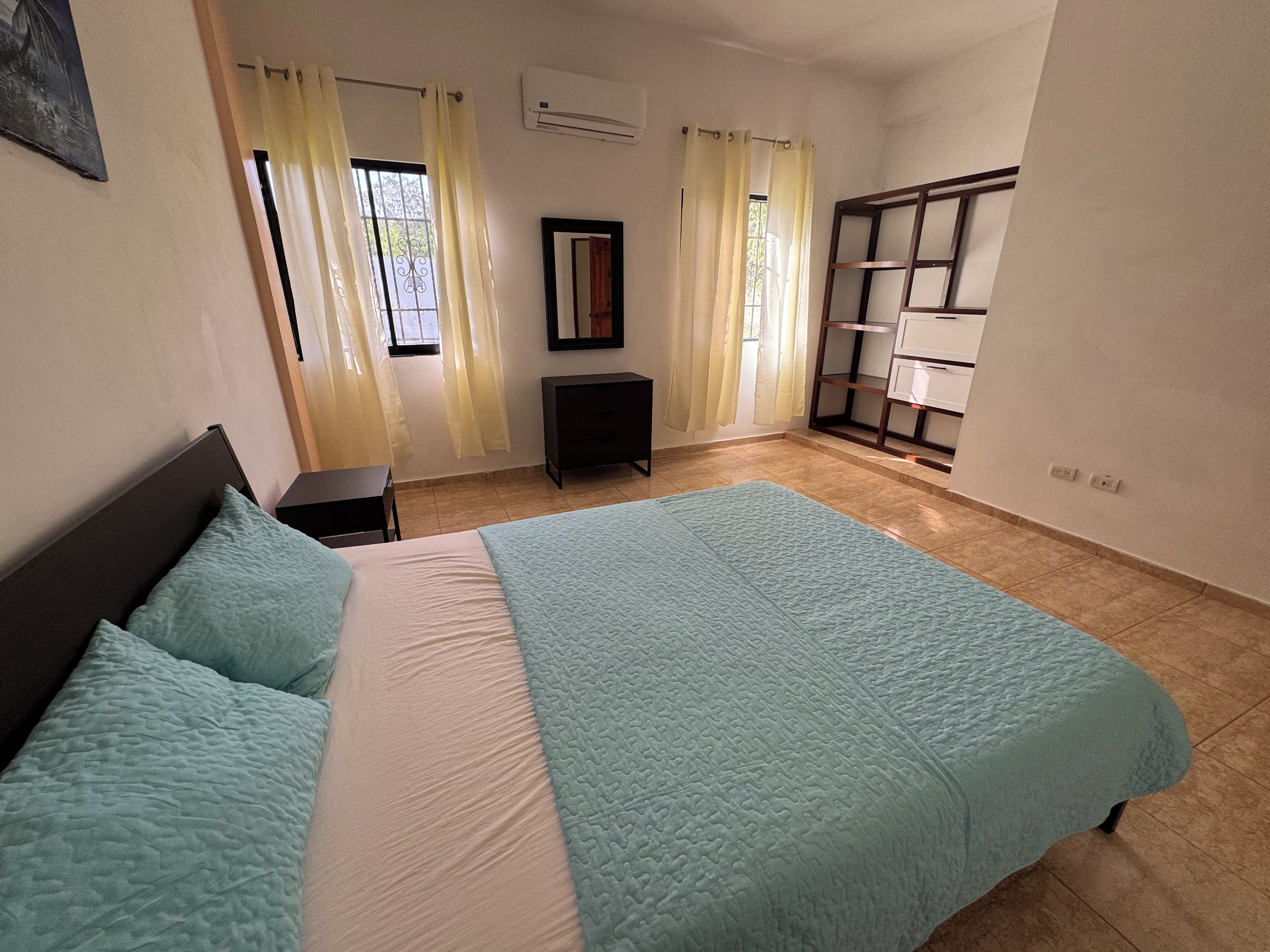 Un dormitorio con cama, mesita de noche, cómoda, espejo y ventana con cortinas.