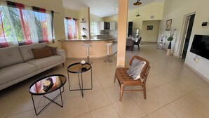 A spacious living area with a sofa, a round coffee table, a bar stool, and a wooden chair. A kitchen is visible in the background with a dining table and chairs. - Casa caleton (Rio San Juan)