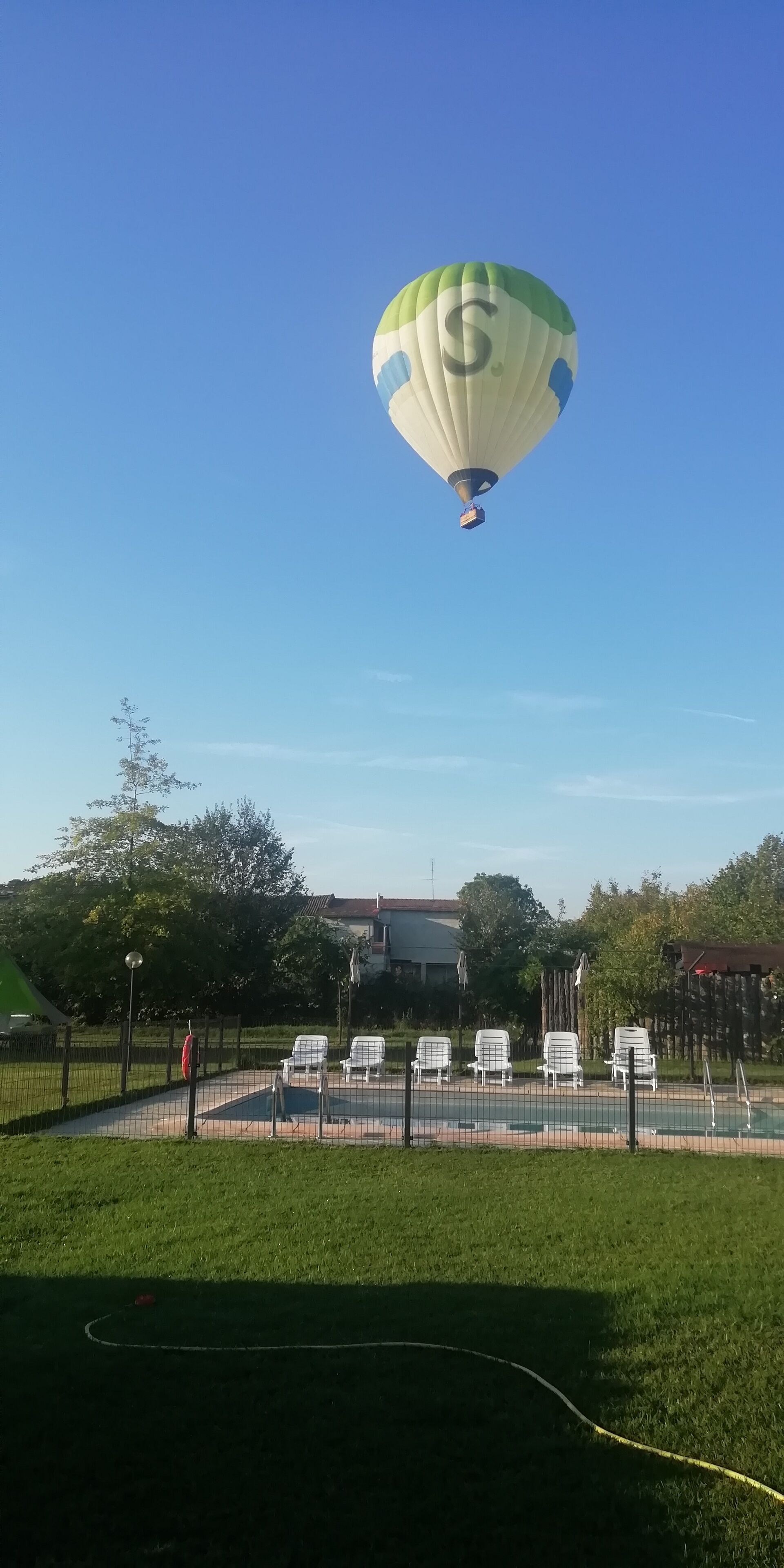 Eine Heißluftballon mit einem 'S' am Himmel, ein Poolbereich mit Umzäunung und Liegestühlen sowie ein Gebäude im Hintergrund.