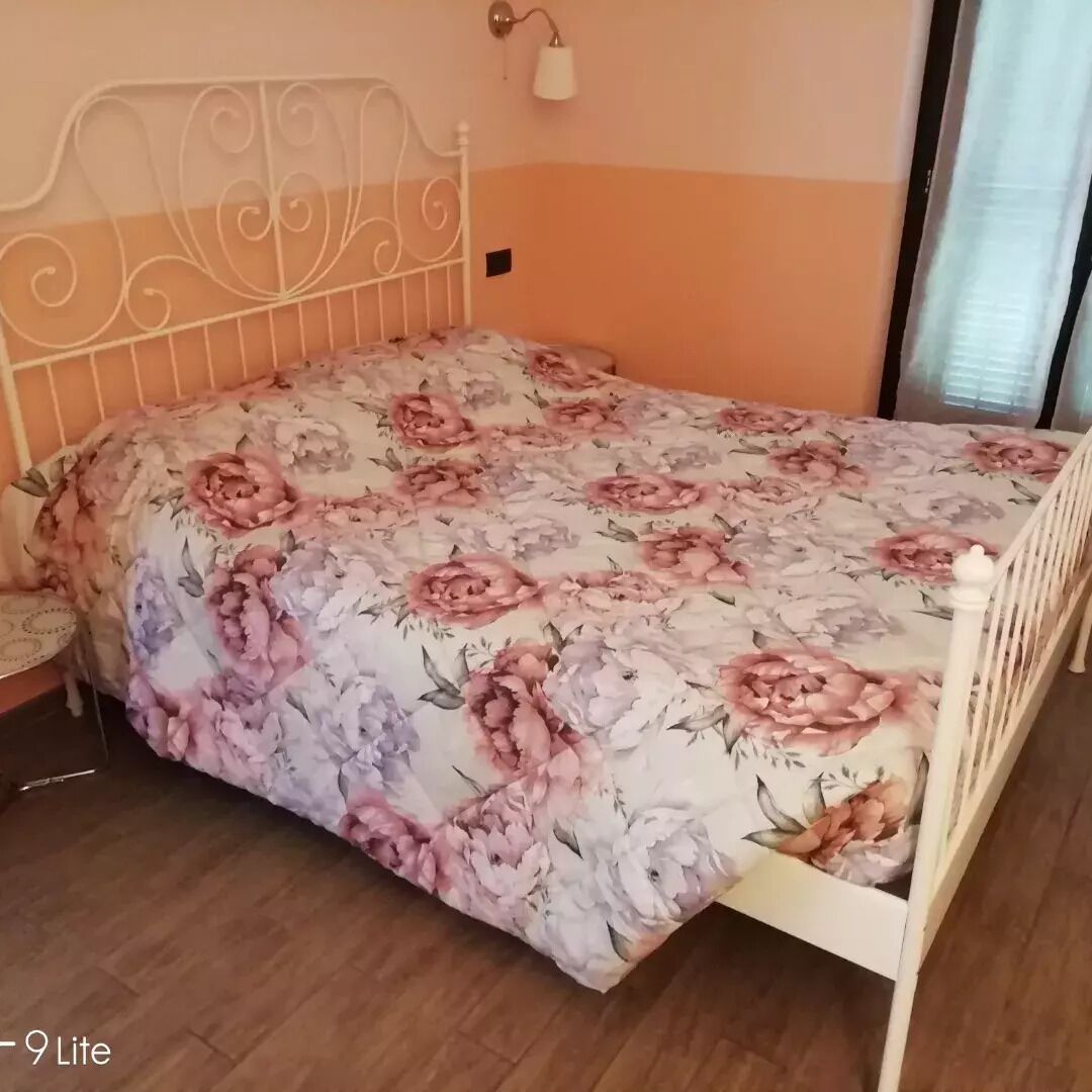 Une chambre à coucher avec une couvre-lit à motifs floraux, un cadre de lit en métal blanc, une table de chevet et une lampe fixée au mur.