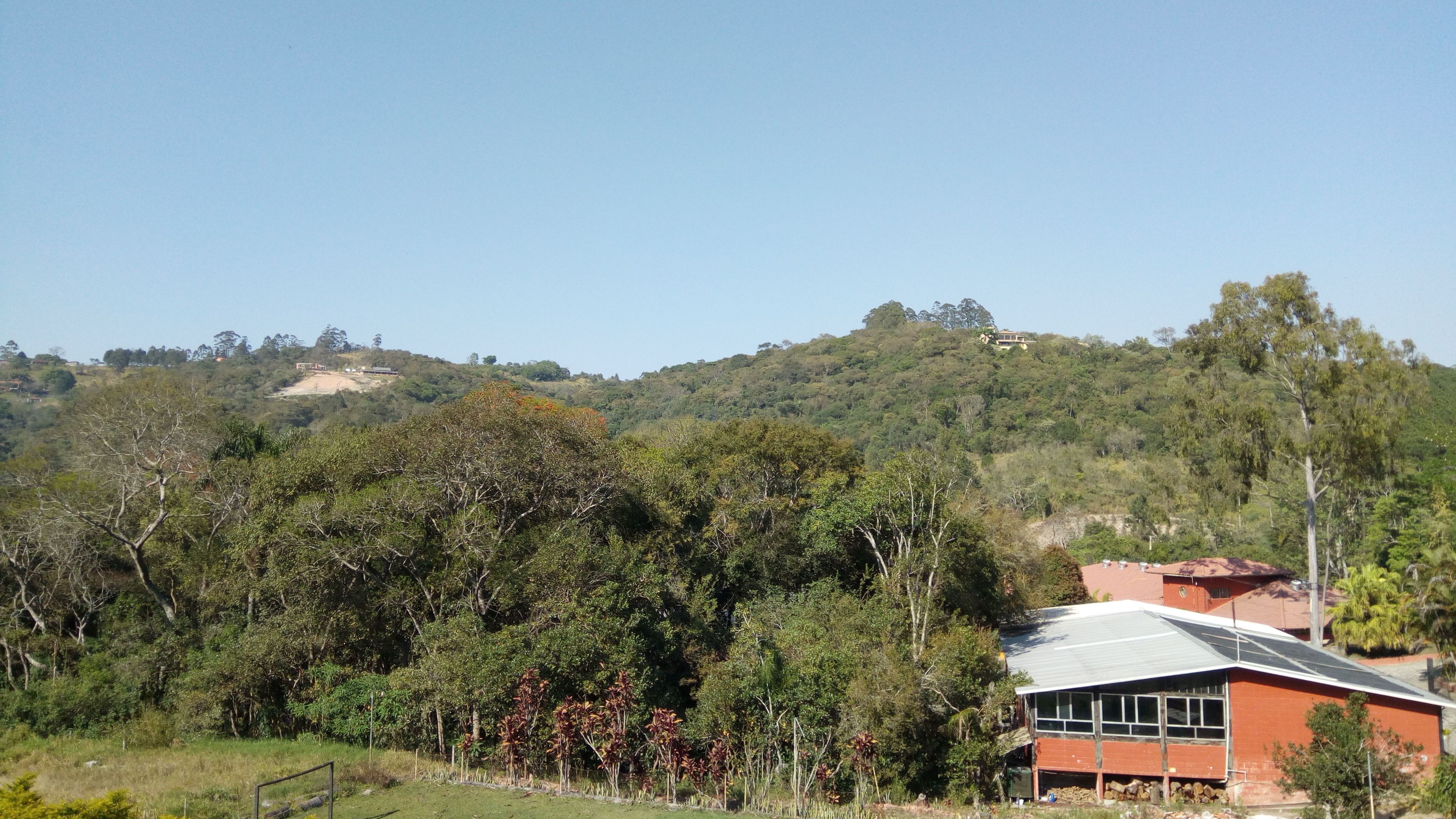 A rural house with a red roof, surrounded by dense vegetation and hills in the background.