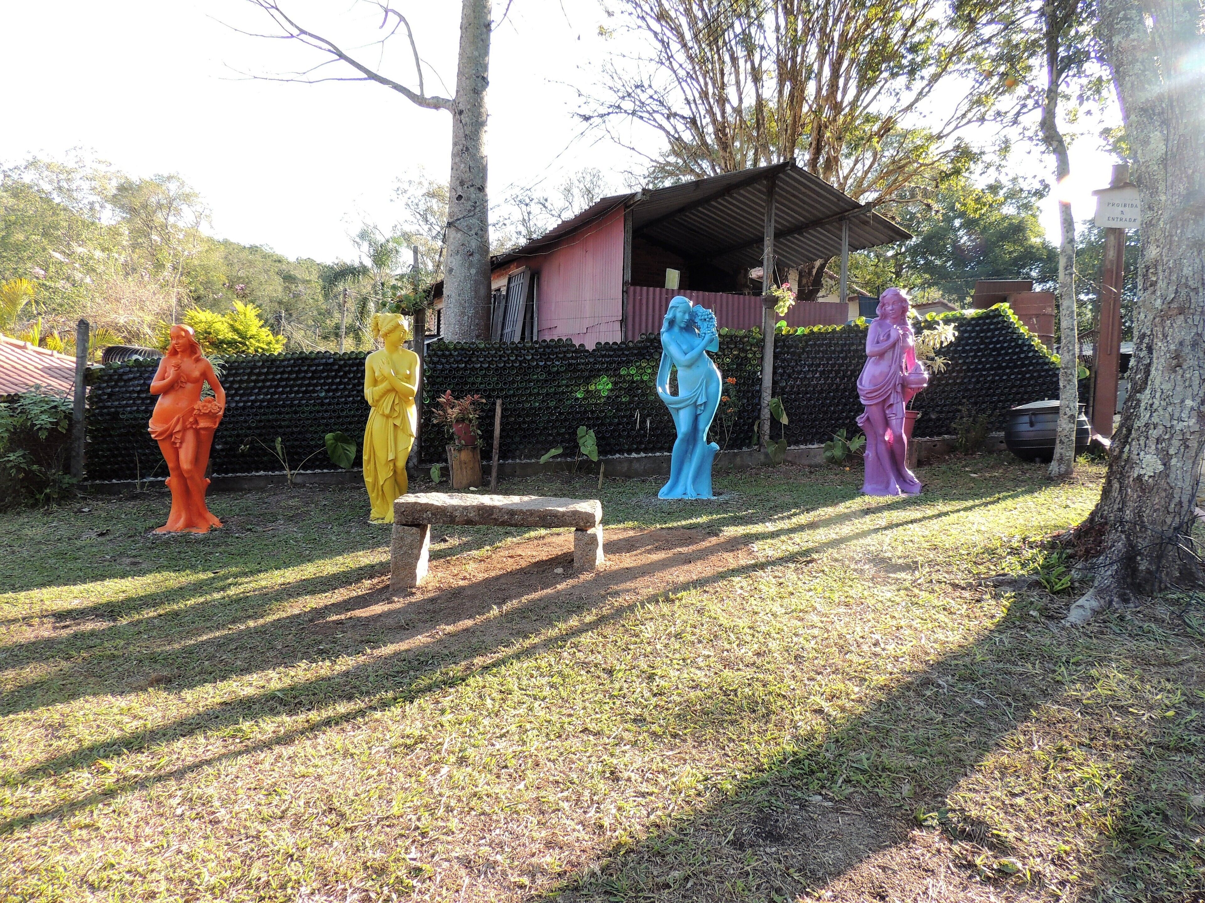 A garden with statues of women in various colors, a wooden bench, and a building with a corrugated metal roof.