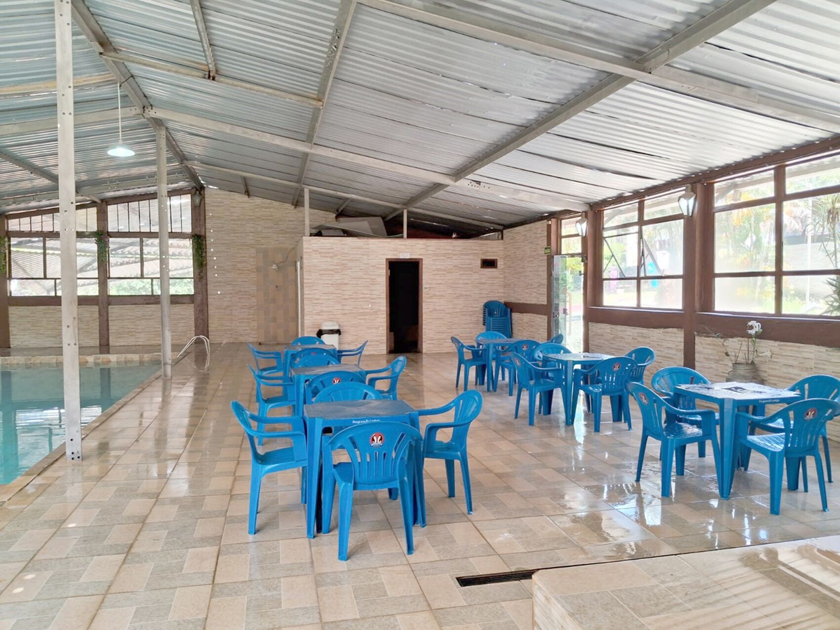 A covered outdoor dining area with blue plastic chairs and tables, a pool, and a brick wall.