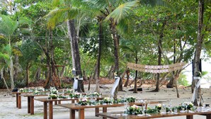 Un área de comedor al aire libre con mesas de madera dispuestas sobre una playa arenosa, rodeada de palmeras y una hamaca.