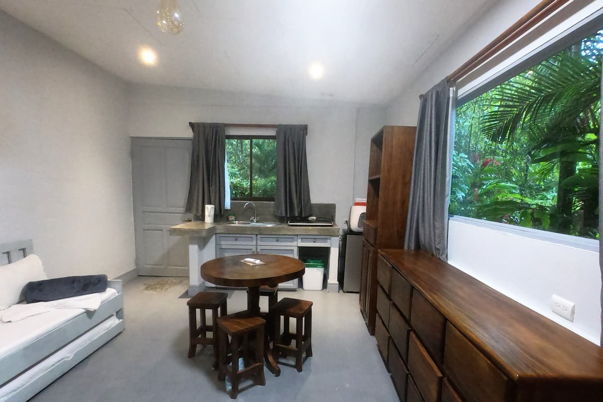 A compact living space with a kitchenette, a dining area with stools, a sofa, and a large window with a view of greenery.