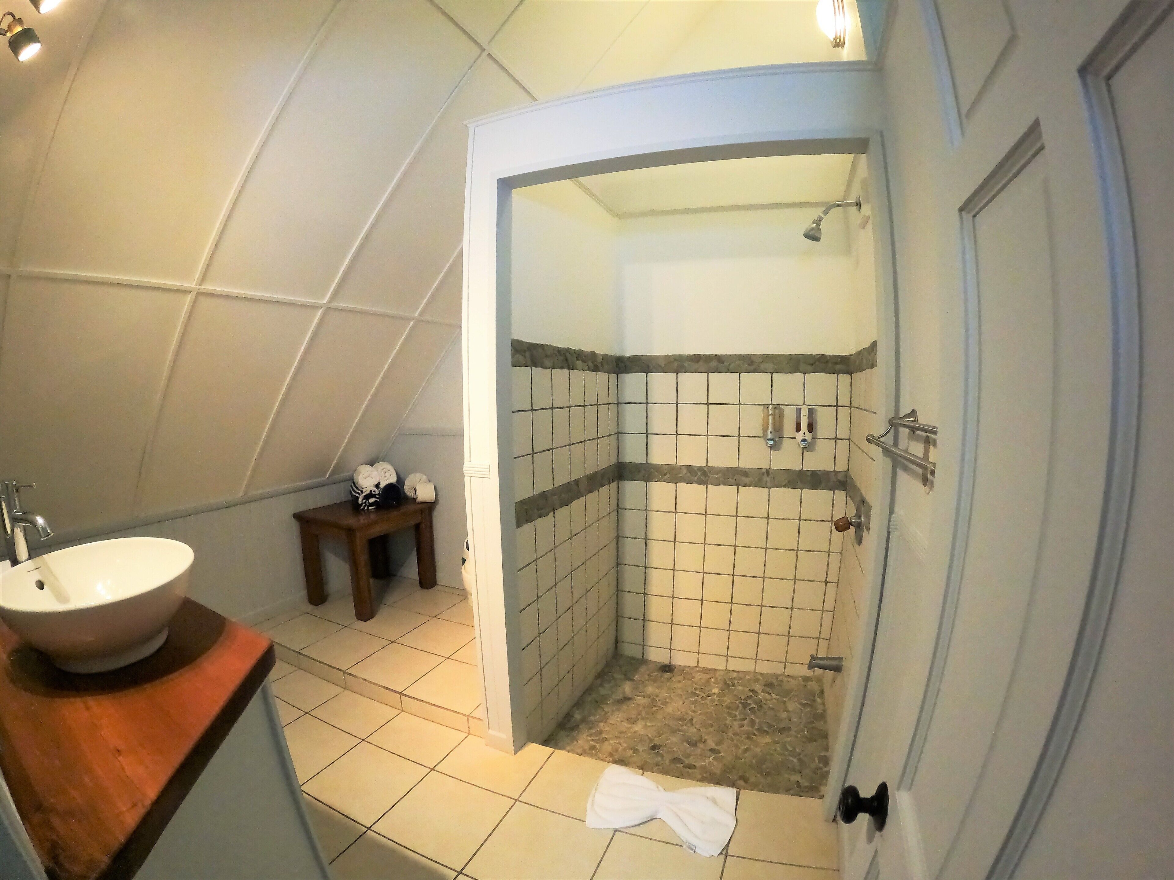 A bathroom with a sink, a small wooden stool, a shower with a tiled wall, and a door.
