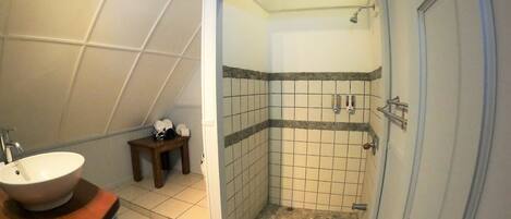 A bathroom with a sink, a small wooden stool, a shower with a tiled wall, and a door.