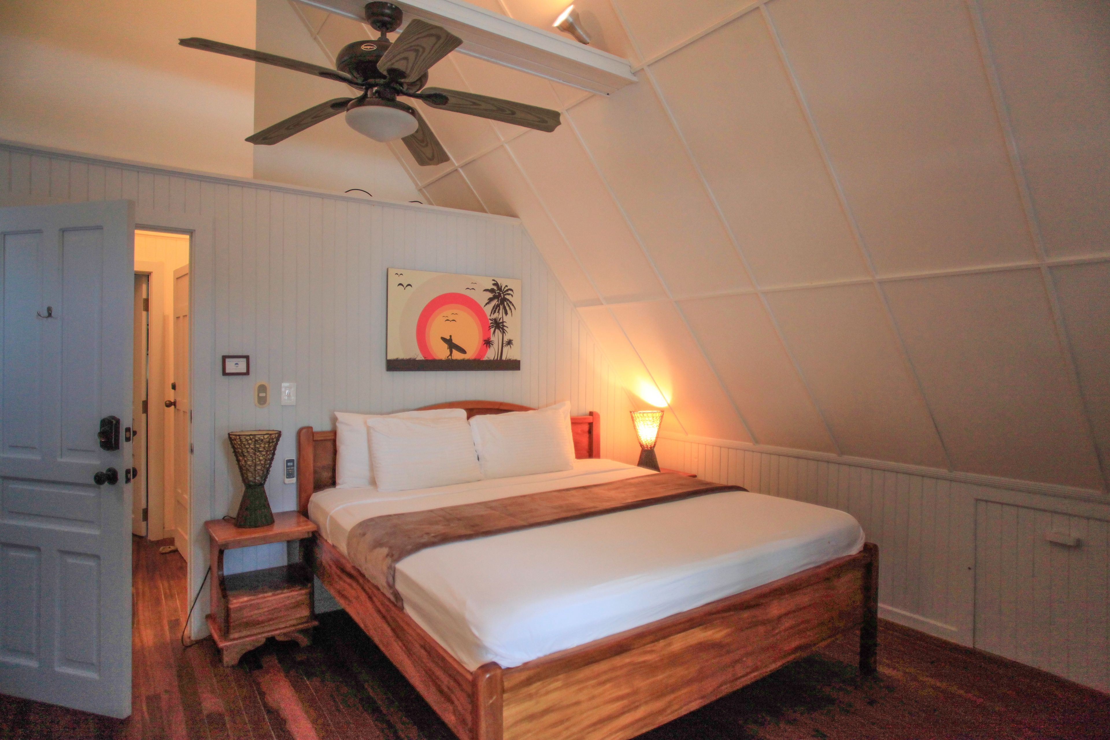 A bedroom with a wooden bed, a ceiling fan, a nightstand, a wall-mounted lamp, and a painting of a sunset.
